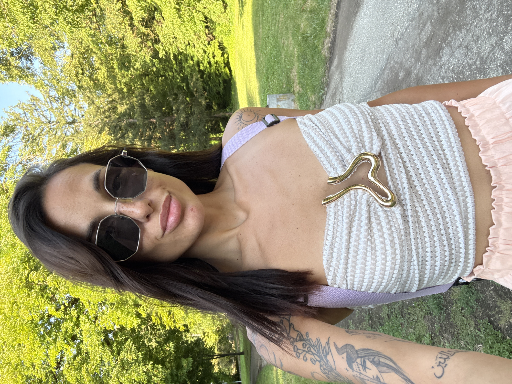 A woman with dark hair and sunglasses is taking a selfie outdoors. She is wearing a beige and white striped tube top with a gold accent piece and a light purple strap. She has tattoos on her left arm and shoulder. She is standing in a park-like setting with green trees and grass. A paved path is visible behind her. The location is Pleven, Bulgaria.