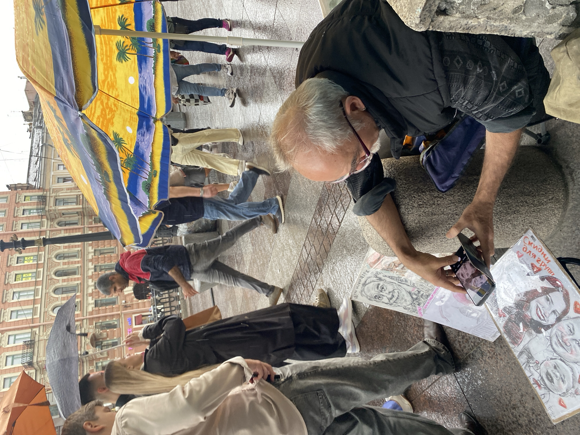 On a rainy day in Saint Petersburg, Russia, an artist sits on the ground, holding a smartphone to photograph his work. He is positioned in front of a display of his drawings, which are laid out on the wet sidewalk. To his right, a large, colorful umbrella shields him from the rain. The backdrop features a classic Saint Petersburg building with its distinctive architectural style. Pedestrians walk by under their own umbrellas, navigating the wet conditions, some of whom are captured in motion. The scene depicts a street artist's resilience and dedication to his craft, even in adverse weather.