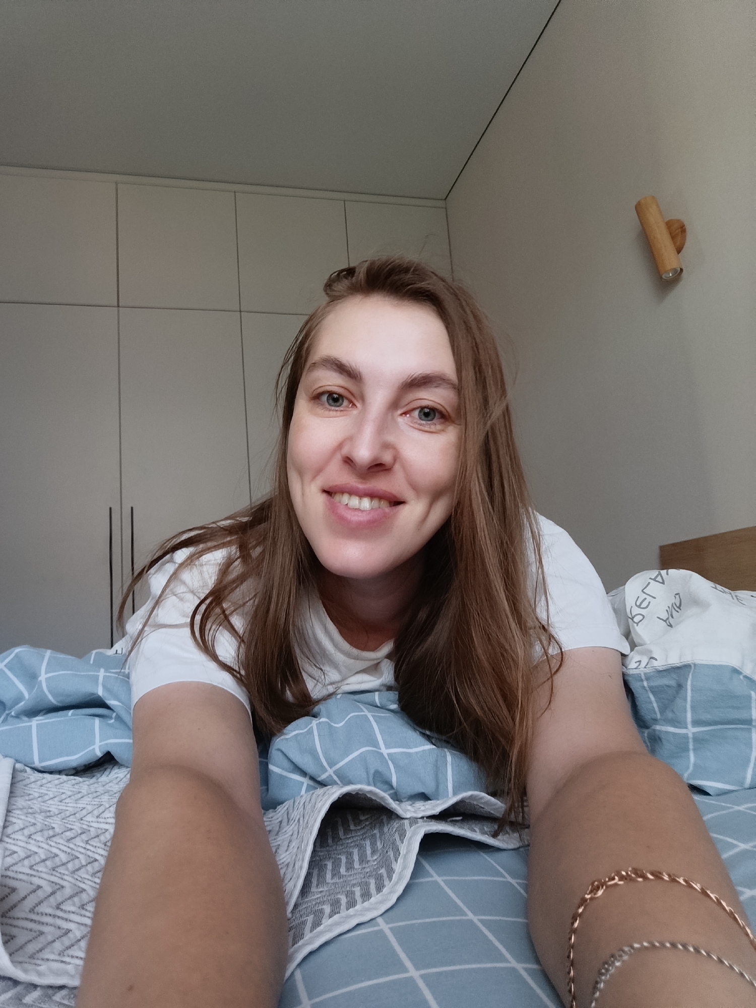 A young woman lies on a bed with blue and white checkered bedding in a room with light-colored walls and built-in cabinetry. She is smiling at the camera. She is wearing a white t-shirt and a bracelet on her right arm. A wooden light fixture is mounted on the wall behind her. Based on the provided information, the location is in the city of Chaikovsky, Russia.