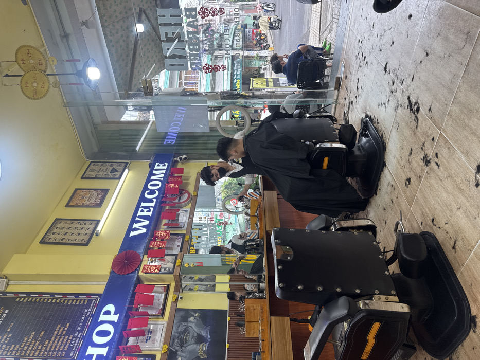 The image displays the interior of a barbershop, identified as "HERO BARBER SHOP" from signage visible on the glass entrance, situated on a street in Nha Trang, Vietnam. The floor is tiled with light-colored squares and is substantially covered with discarded strands of cut hair.
In the foreground, an empty, black, reclinable barber chair with chrome and gold accents faces away from the camera. Directly behind it, a male barber, wearing a black vest over a white shirt and a black face mask, is actively cutting the hair of a male customer. The customer is seated in a similar black barber chair, draped in a black cape, with his back to the camera. The barber holds clippers in his right hand, positioned near the customer's head.
The shop's interior features a yellow upper wall and a lower section with wooden paneling. A prominent blue banner displays the illuminated word "WELCOME" in white letters, beneath which numerous red envelopes and a circular red paper fan are suspended. A framed price list in Vietnamese, along with other framed pictures and a large black-and-white mural, adorn the walls.
Through the clear glass storefront, a street scene characteristic of Nha Trang is visible, featuring multi-story buildings, parked and moving motorbikes, and several individuals seated on small stools outside the shop entrance. Reflections on the glass show elements of the interior and the reversed text "HERO BARBER SHOP." Overhead, fluorescent lighting and two decorative circular yellow hangings are visible.