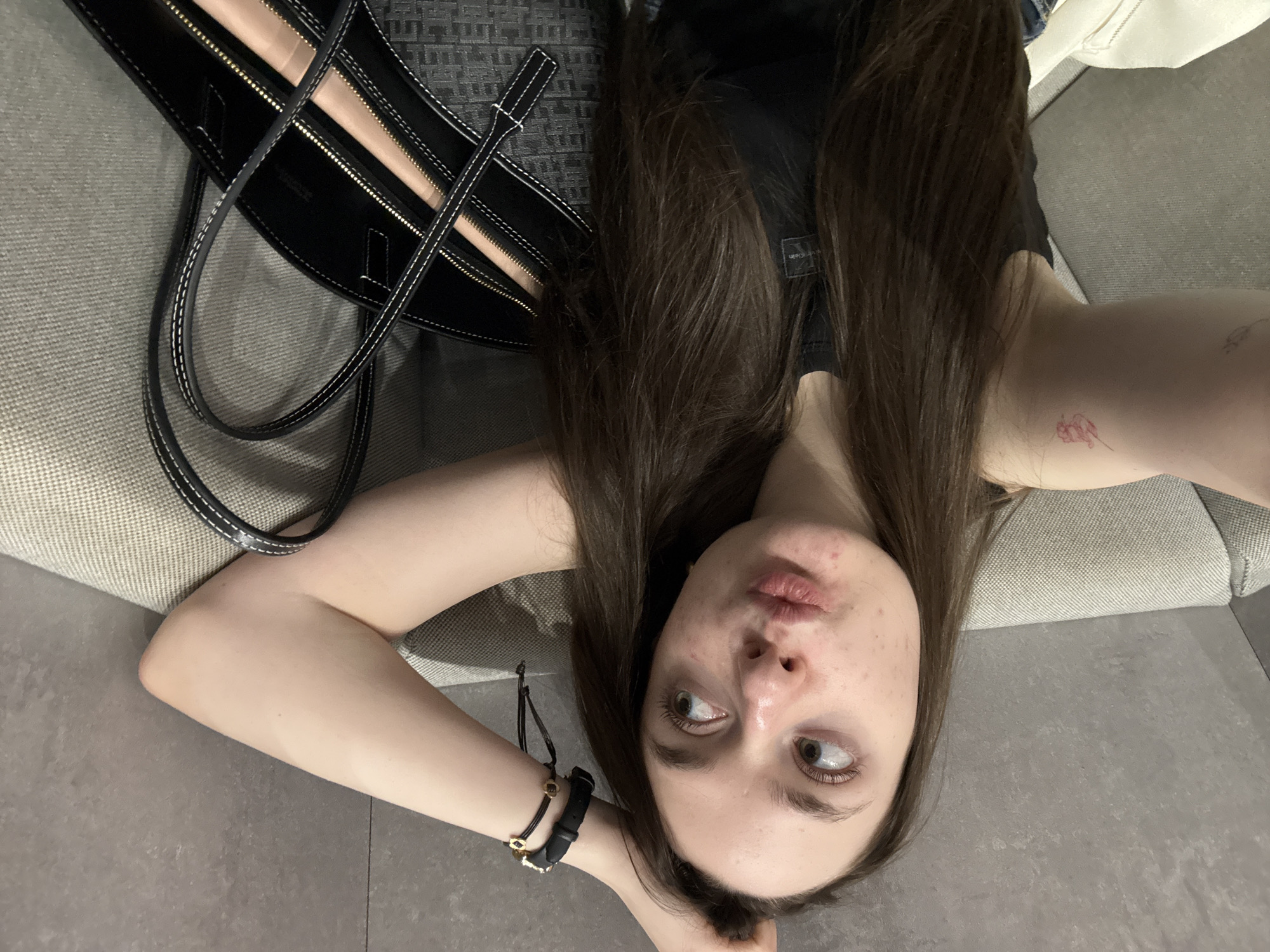 A woman with long brown hair is reclining on a gray upholstered surface, possibly a sofa or bed. She is angled towards the camera, her head slightly tilted. She has fair skin and noticeable acne on her face. She is wearing a black tank top and has a wristband on her left wrist. A black handbag with light-colored lining and white stitching is positioned near her head. The location of this image cannot be reliably determined to be in Fiumicino, Italy, based solely on the visual information provided.