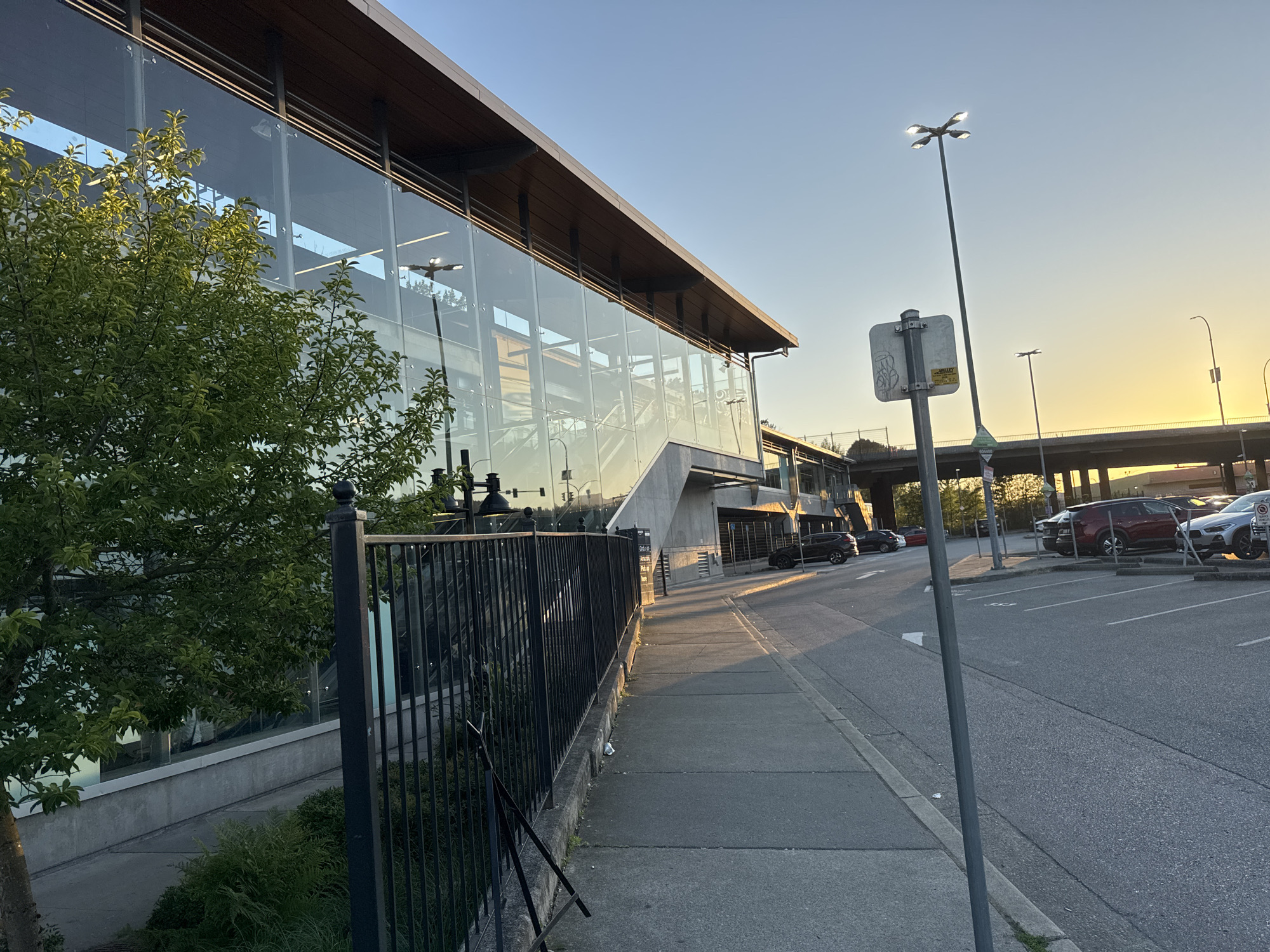 This is an outdoor, eye-level, horizontal shot of a modern building with a glass facade on the left, adjacent to a sidewalk and a parking lot. The building appears to be a public transit station, possibly a SkyTrain station, given the architecture. The sun is setting, casting a warm golden light on the scene and creating long shadows.
In the foreground, a sidewalk runs along the right side of a black metal fence that borders the building. Green foliage is visible behind the fence and in front of the glass facade. The sidewalk is made of concrete and shows some wear.
To the right of the sidewalk is a paved parking lot with several cars parked. The cars are of various colors and models, including a dark-colored SUV, a red SUV, and a silver SUV. The parking lot is marked with white lines. A tall streetlight with multiple heads stands in the middle of the parking lot, casting a bright light. A street sign with graffiti is attached to the pole of this streetlight.
In the background, a highway overpass is visible, with traffic flowing on it. The sky is a clear blue, transitioning to a golden yellow near the horizon, indicating sunset. The overall scene depicts an urban environment at dusk, likely in Coquitlam, Canada, given the characteristic SkyTrain architecture and general urban landscape. There are no people visible in the image.