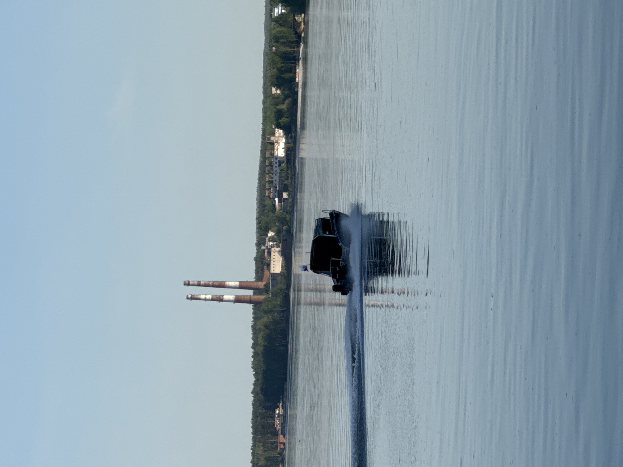 The image shows a motorboat speeding across a body of water, likely a river or a large lake, in Chaikovsky, Russia. The boat is trailing a visible wake. On the shoreline, a dense line of trees and structures are visible. Two tall industrial chimneys, banded in red and white, rise from the landscape. The sky is clear and light blue.
