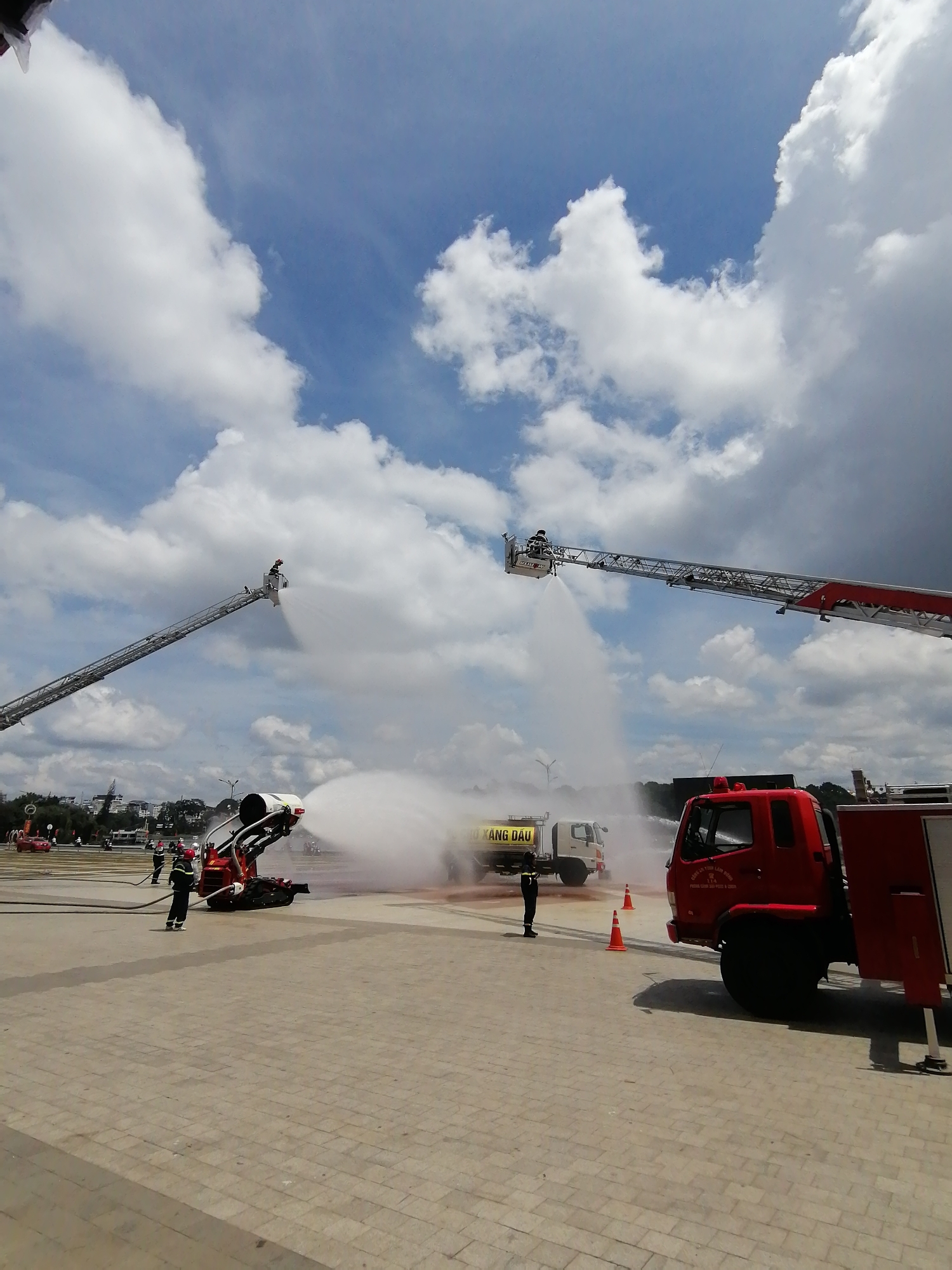In the city of Da Lat, Vietnam, two fire truck ladders extend upwards, spraying water into the sky, creating a misty effect. One ladder is to the left, with a firefighter in a bucket at its tip. The other ladder is to the right, also with a firefighter at its tip. In the foreground, a red fire truck is parked. Behind it, a white truck with "XĂNG DẦU" written on its side is visible. Several firefighters in uniform are present on the ground. The scene unfolds on a paved, open area under a partly cloudy sky.