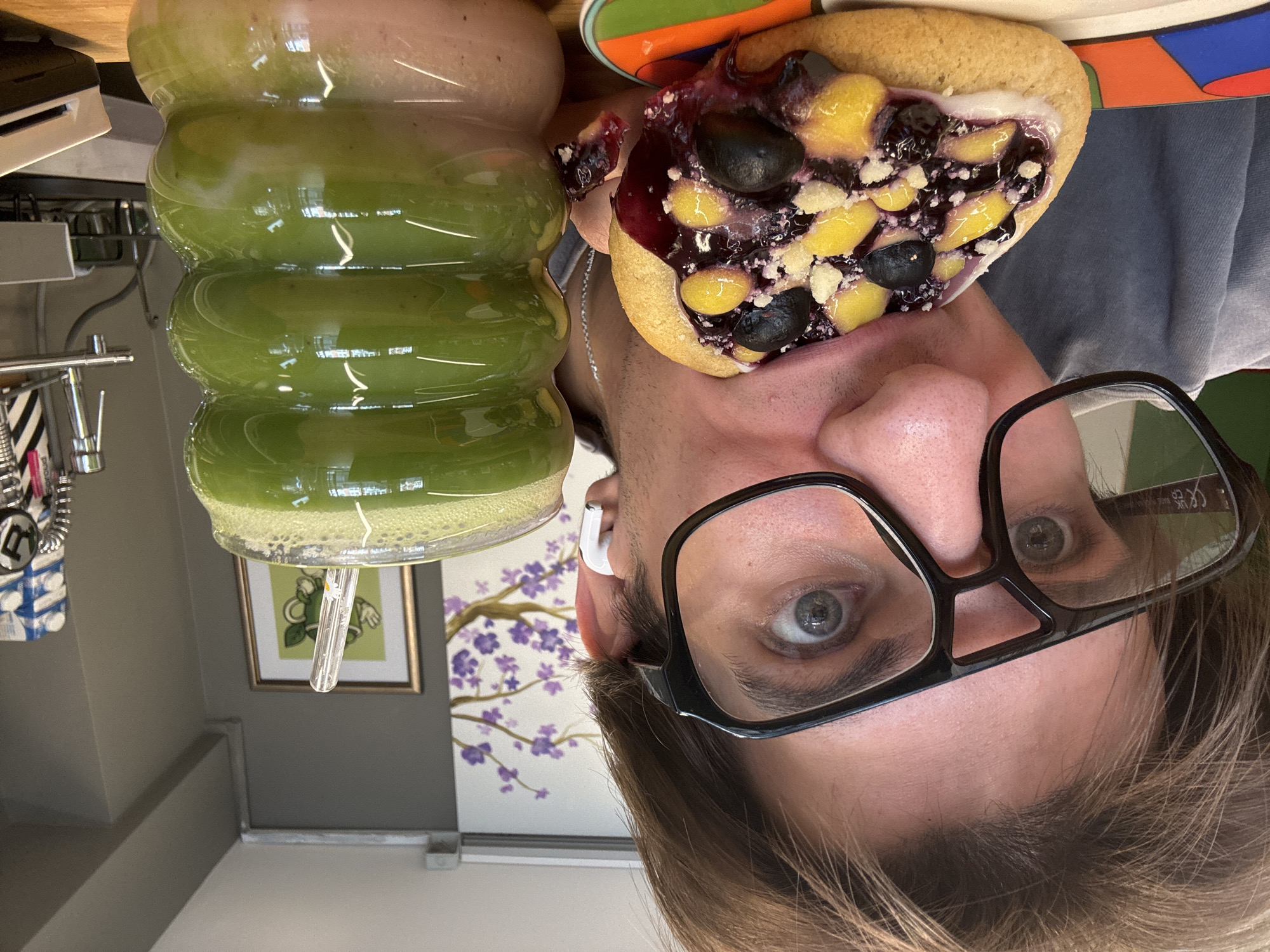 The image is an upside-down close-up selfie of a person with light skin, light brown hair, and gray eyes, wearing black rectangular glasses and a white wireless earbud in their left ear. The person is holding a heart-shaped cookie topped with blueberries, yellow fruit chunks, and white crumble topping next to their face. A glass with a green smoothie is positioned next to the cookie. The background includes part of a countertop, a wall painted gray on top and white on the bottom, and two framed artworks – one featuring a green cartoon character, and another with purple flowers. Due to the lack of discernible elements, the location cannot be determined as Sofia, Bulgaria.
