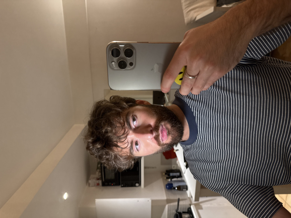 A male individual with brown, curly hair and a beard is depicted taking a mirror selfie indoors. He wears a short-sleeved t-shirt with horizontal dark blue and white stripes and a dark blue collar. His left hand, adorned with a silver-colored ring on the ring finger, holds a silver/gray Apple iPhone with three rear camera lenses. A yellow circular sticker featuring a black smiley face is affixed to the back of the phone. The individual's gaze is directed slightly to his right, towards the reflection.
The background on the left side of the frame reveals a kitchenette area, including white upper and lower cabinets, a light-colored countertop, a small black coffee maker, several dark bottles, a microwave oven, and another black appliance stacked beneath the microwave. Recessed lighting is visible in the ceiling. The background on the right side of the frame shows a plain light-colored wall and a partial view of a white upholstered item, possibly a headboard or sofa. The specific location within the city of Paris, France, is not discernible from the visual elements present in the image.