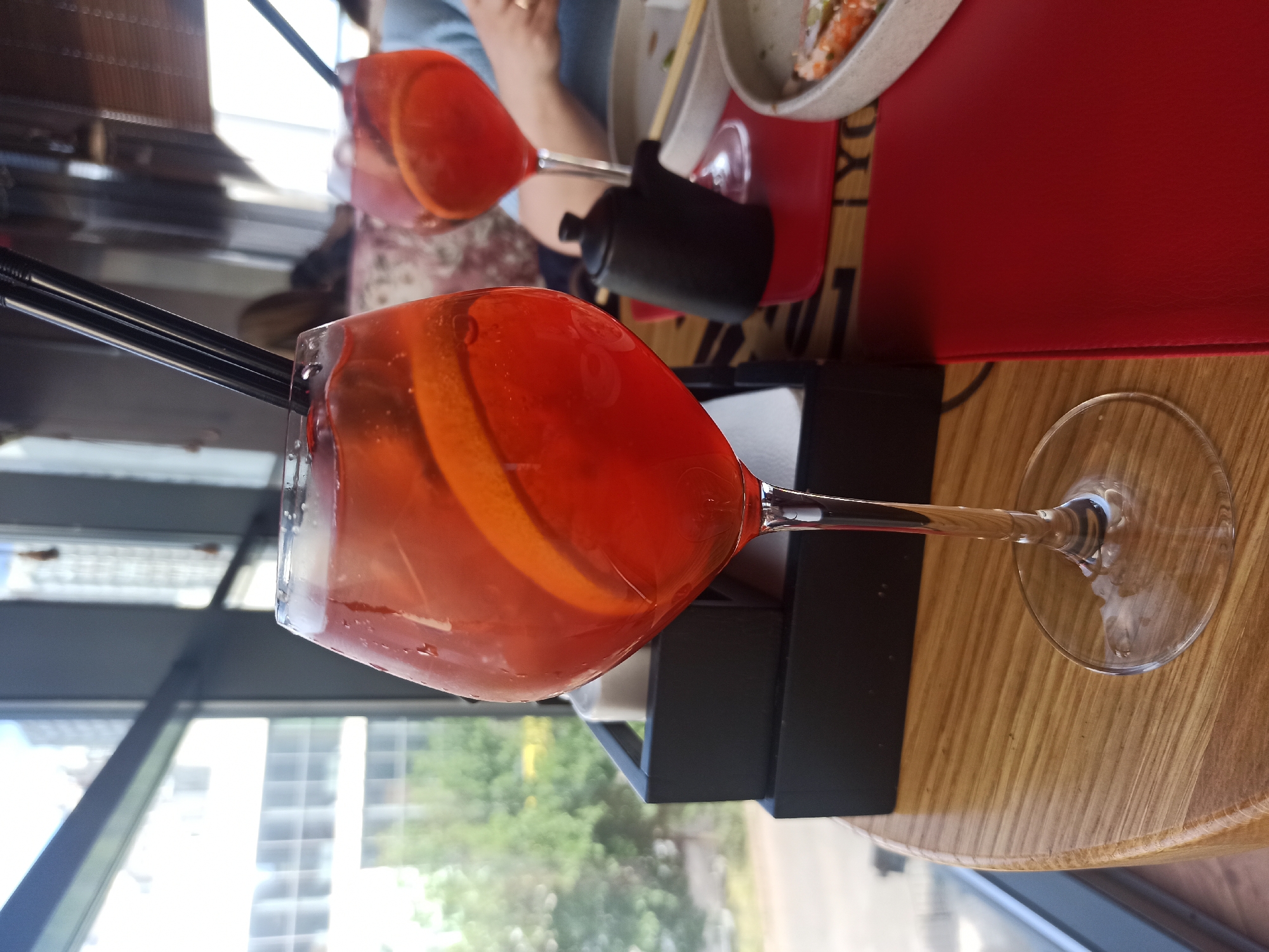 The image depicts a close-up, tilted shot of two Aperol Spritz cocktails served in stemmed glasses. The primary cocktail, in focus, has a reddish-orange hue and includes a slice of orange, with two black straws protruding from the glass. It sits on a black stand placed on a wooden table.
In the background, partially out of focus, is another Aperol Spritz cocktail. To the right of this cocktail is a black container, potentially for soy sauce, and a dish with food items. A red menu or placemat is also visible.
The scene is set next to a window with a metal frame, looking out onto an urban environment with trees and buildings. The location appears to be indoors, likely a restaurant or bar.