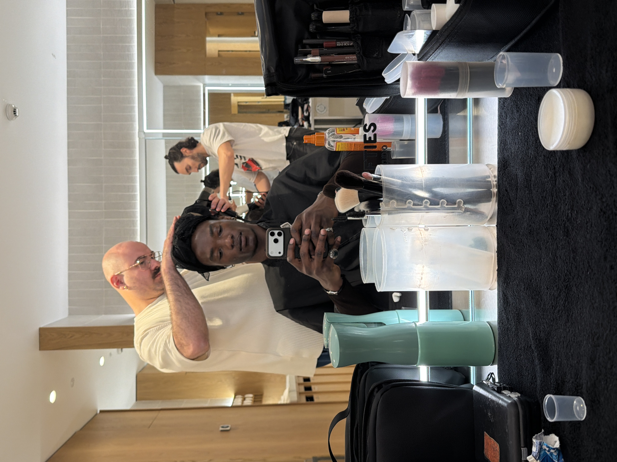 The scene takes place indoors, possibly in a beauty salon or studio. Three people are visible: a bald man in a white shirt standing on the left, touching the hair of a person in a black robe taking a selfie; and another man in a white shirt working with another person in the background. The selfie taker is holding a smartphone. Several makeup brushes and tools are visible on a black surface, along with containers, cases, and a spray bottle, suggesting a professional makeup setup. The room appears modern, with white walls and light wood accents. The image does not give location information to confirm whether this is in the city of Fibracolor, Spain.