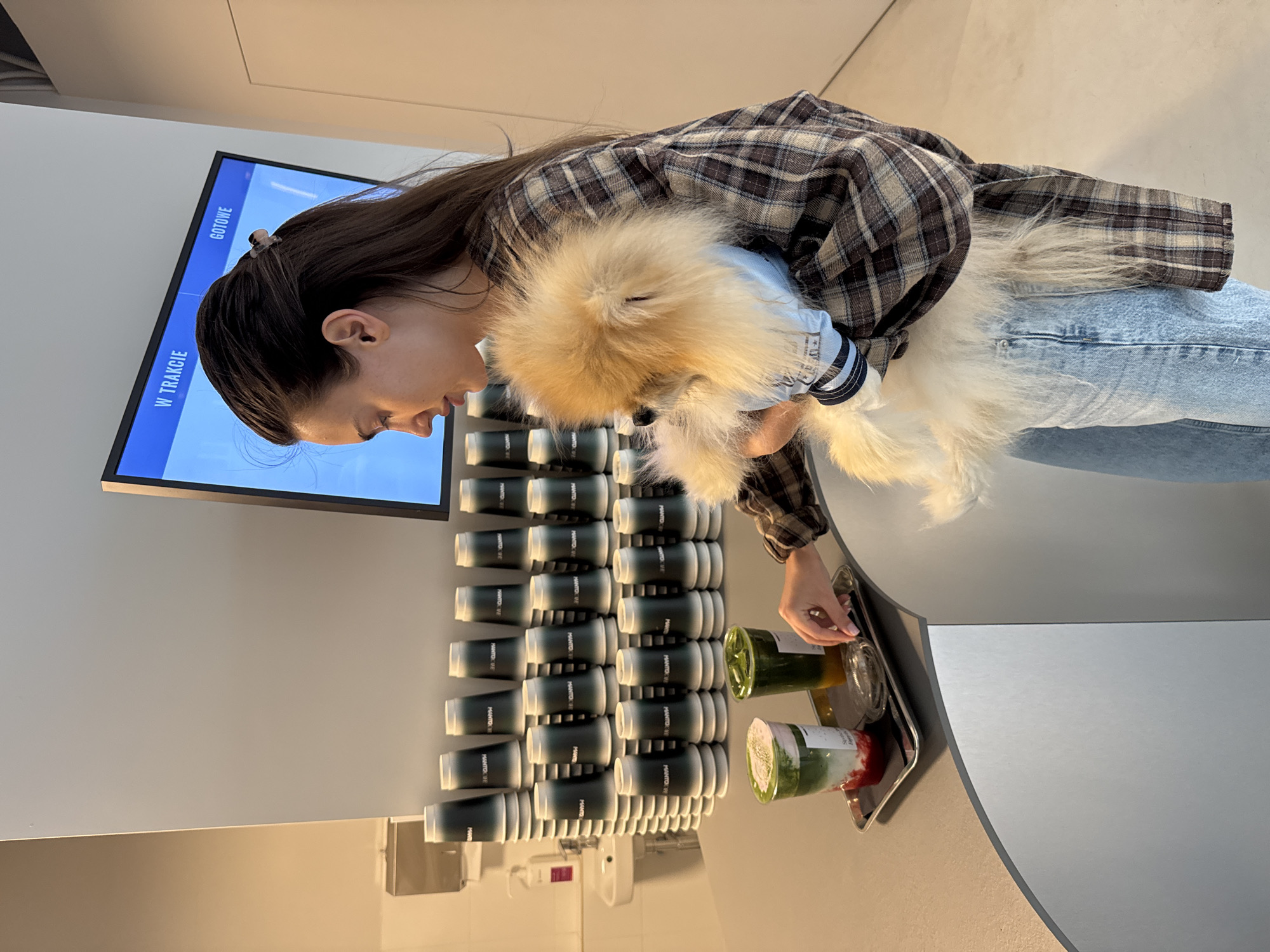 An indoor scene features a young woman at a counter, holding a small, fluffy dog. The image is presented in a vertical orientation. The woman wears a plaid shirt in shades of brown, white, and black, paired with light blue jeans. Her dark hair is pulled back, revealing a small clip. She gently holds a cream-colored, fluffy dog, possibly a Pomeranian, which is wearing a light blue top with white striped sleeves.
On a silver tray resting on the light grey counter, there are two clear plastic cups containing layered beverages; one is green and yellow, while the other is green, white, and reddish-pink. The woman's right hand is near the drinks, possibly adjusting a lid or selecting a straw.
Behind her, a wall features a display of numerous dark green/grey cups, neatly stacked in multiple rows. Each visible cup is printed with the text "PANTO VR." To the woman's left, a flat-screen monitor is mounted on the wall, displaying text in a light blue interface. The visible text reads "W TRAKCIE" (Polish for "In Progress") and "GOTOWE" (Polish for "Ready"), suggesting an order status display. A white dispenser is visible on the wall beneath the stacked cups. The setting appears to be a modern cafe or juice bar. The woman has a calm and gentle expression as she looks down, likely at her pet or the drinks.