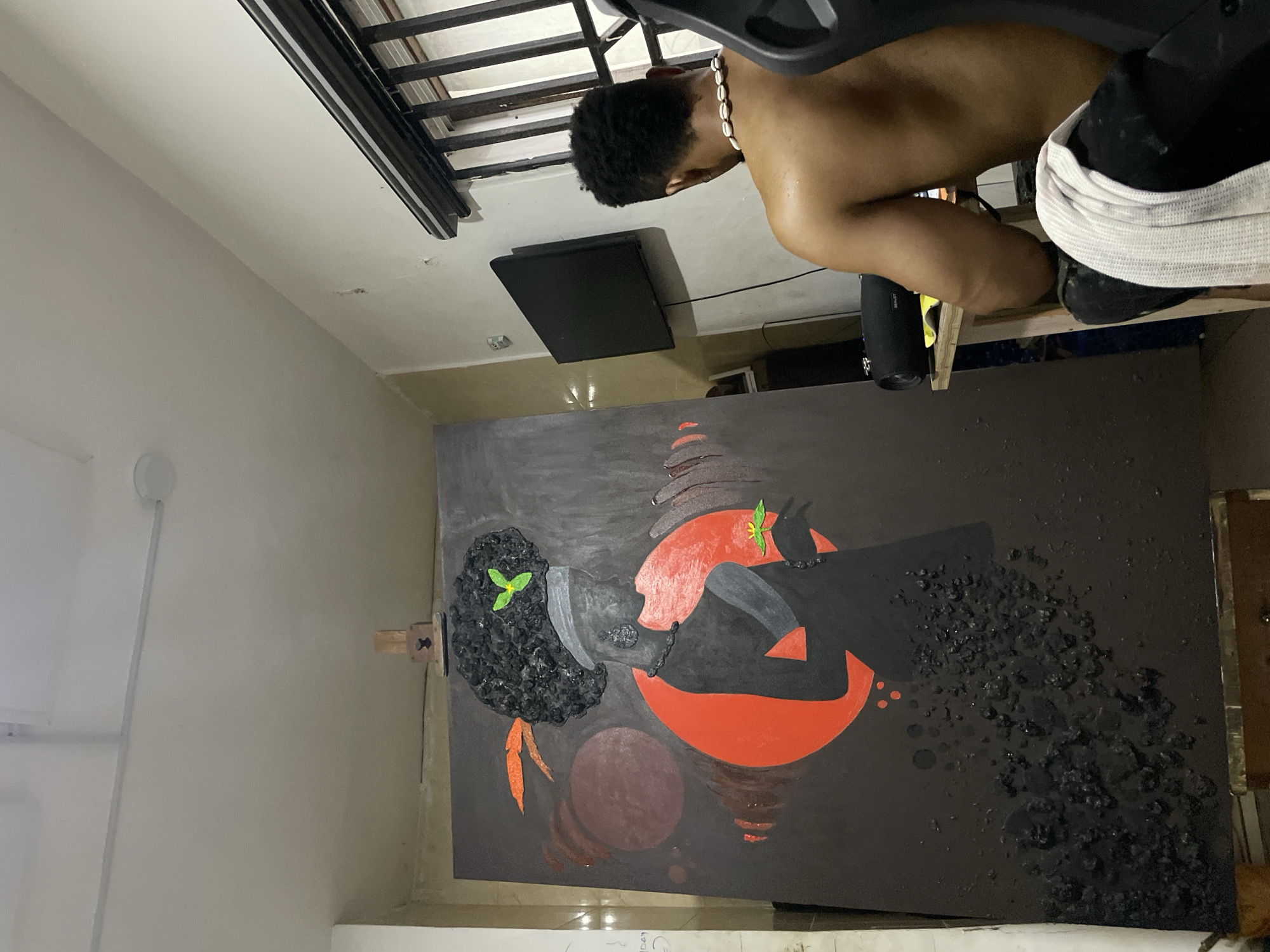 A shirtless man with a beaded necklace is working on a large, abstract painting in an indoor setting. The painting features two figures in black, with textured, three-dimensional elements, set against a dark background with red and brown circular shapes. The setting appears to be a domestic space in Ado, Nigeria, as implied by the prompt. The man is positioned near the painting, suggesting he is the artist.