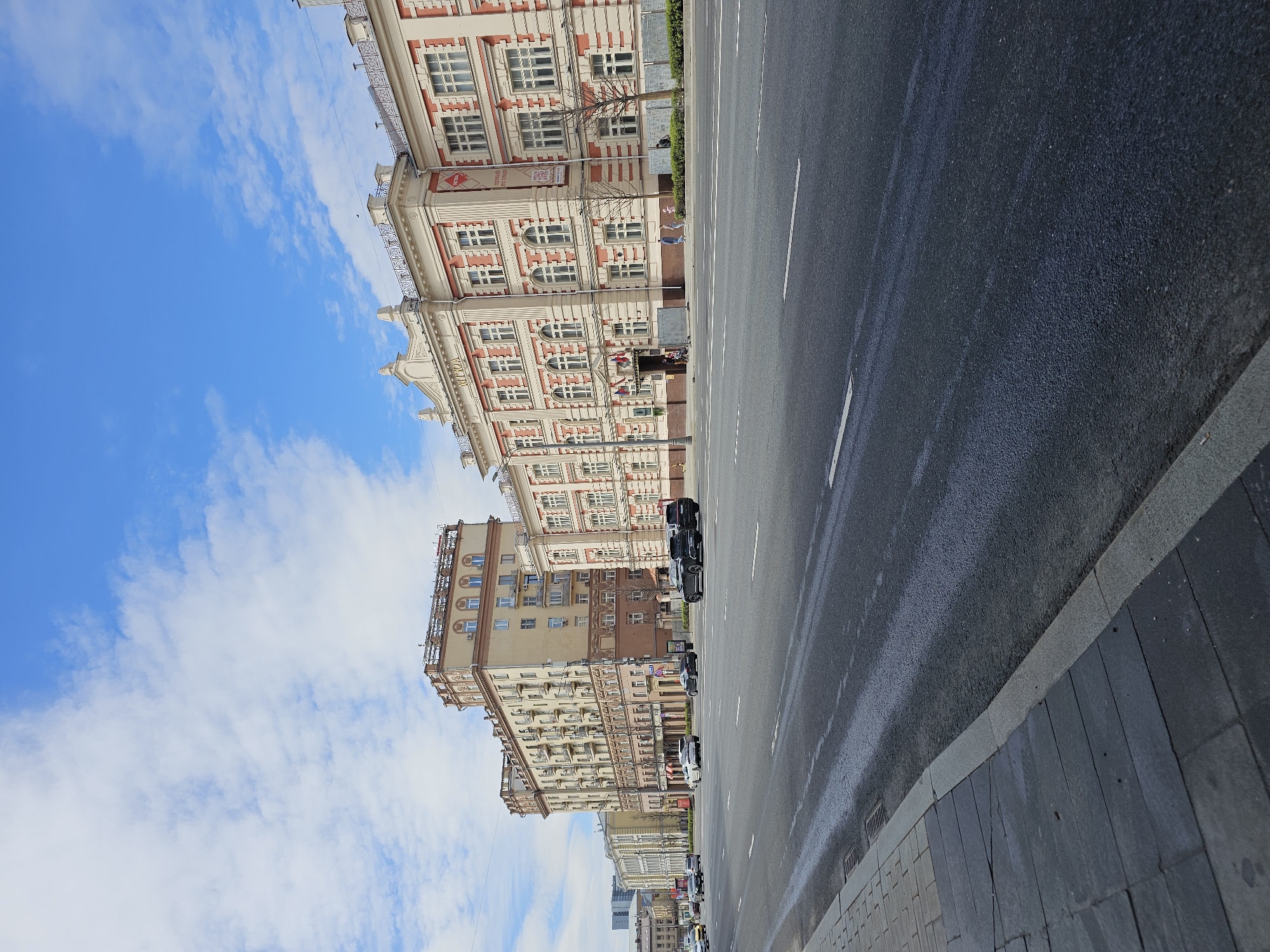 A ground-level, outdoor, long shot depicts buildings lining a street in Moscow, Russia. The camera angle is tilted slightly to the left. The sky is blue with scattered white clouds. Buildings of varying heights are positioned along the left side of the road. The buildings feature architectural details with windows, balconies, and ornate decorations. The foreground consists of a dark asphalt road with white lane markings and a concrete sidewalk lined with stone tiles. Several parked cars are visible along the road's edge. The lighting suggests daylight conditions.
