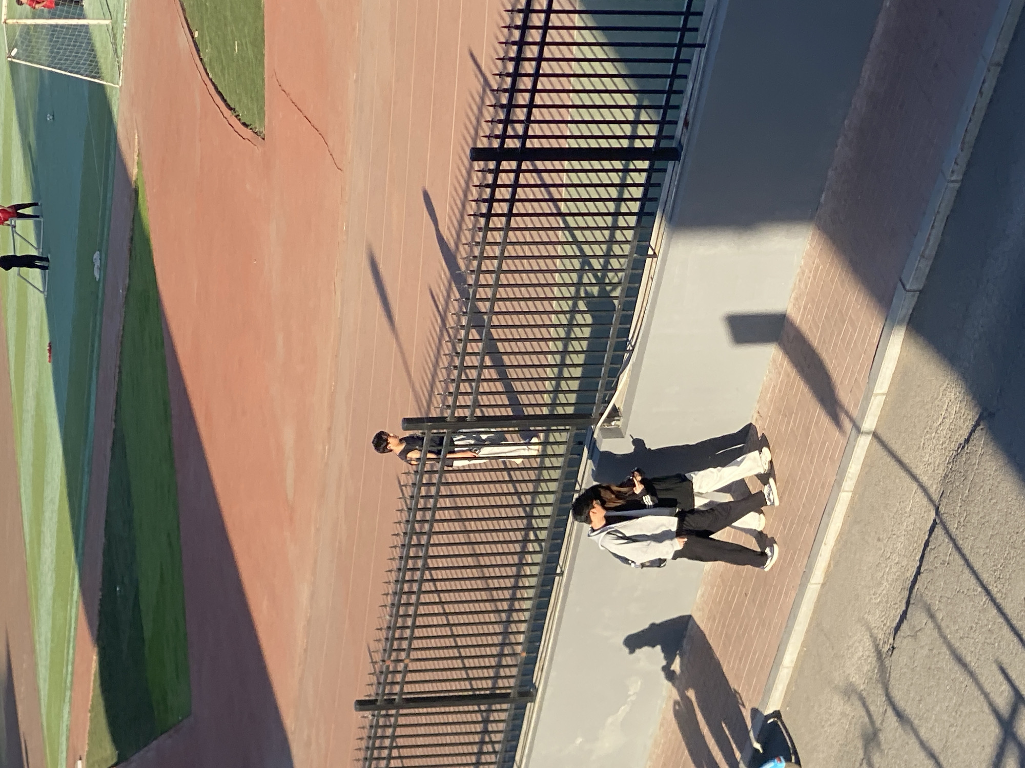 A high-angle, slightly tilted photograph captures an outdoor scene in Zhongtun, China, featuring a sports field and a walkway separated by a black metal fence. The sports field has a grassy area and a running track, with visible goalposts. Two people stand near the fence, looking out over the field. One person is wearing a light-colored jacket and dark pants, and the other is wearing a dark top and light pants. A third person is further away, near the fence, wearing a dark shirt and light pants. The walkway is made of light-colored pavers with a gray concrete edge, leading to a paved road area. Shadows are prominent, suggesting strong sunlight. The overall composition is captured from a raised vantage point.