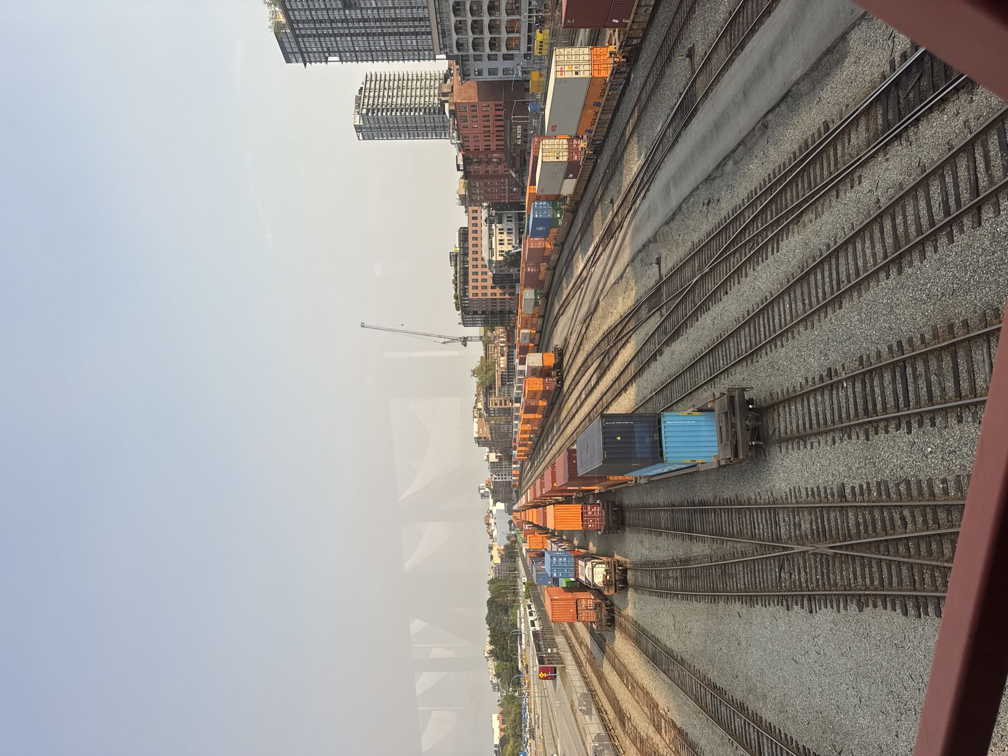 The image shows a train yard with multiple sets of railway tracks in Vancouver, Canada. Several freight trains loaded with shipping containers are visible on the tracks. The containers are of various colors, including orange, blue, and white. In the background, city buildings and a construction crane can be seen. The sky is clear and the overall lighting suggests it is daytime. The perspective is from an elevated position, looking down on the train yard.