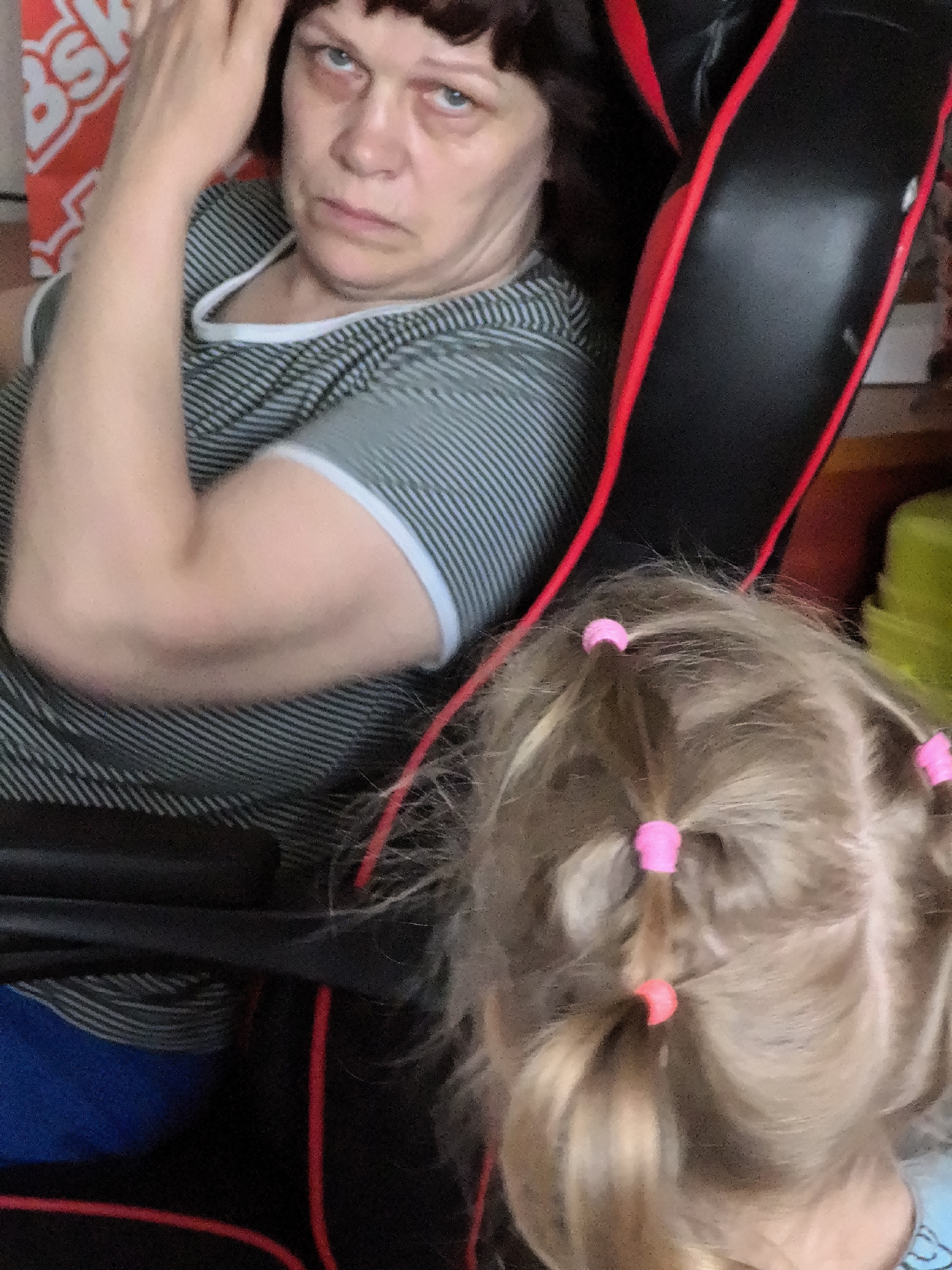 In the foreground, the back of a child's head is visible, with blonde hair styled into several small ponytails secured with pink hair ties. The child is facing away from the camera. Behind the child, an adult woman with dark hair and a striped shirt is seated in a black and red gaming chair. Her left arm is raised to her head, and her gaze is directed forward with a stern expression. The background is blurred, but a red and white patterned object is partially visible to the left, and a green object is visible to the right. The scene appears to be indoors, possibly a home environment.