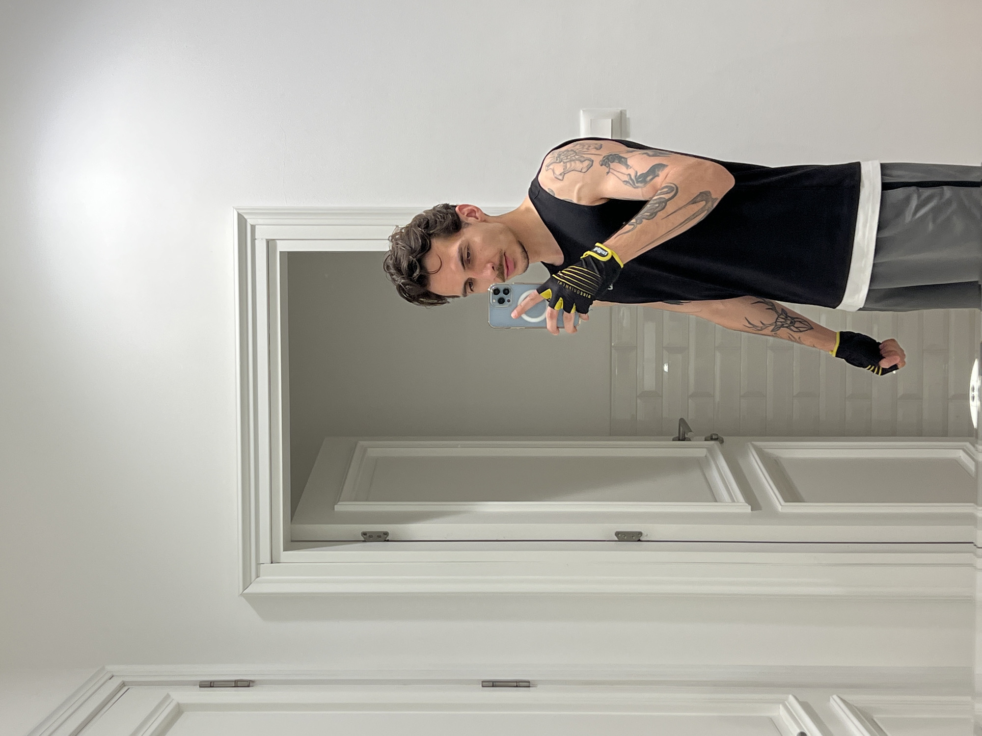 A man stands in front of a mirror taking a selfie. He is wearing a black tank top, grey shorts, and black and yellow gloves. He is holding a light blue phone. The mirror reflects a white tiled wall and a door with white frames. The setting appears to be a bathroom. There is no information to determine the location in the city of Boreti, Montenegro.