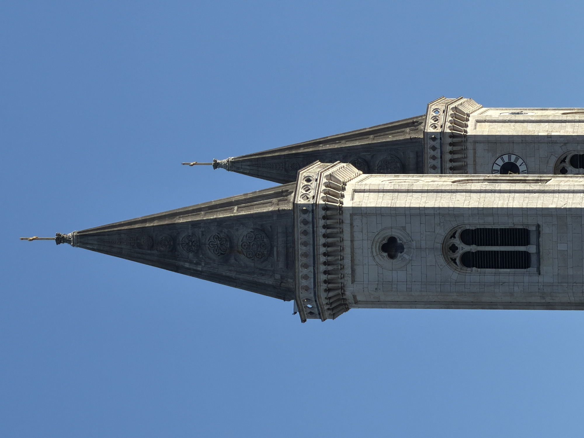 The image shows two spires of a cathedral against a clear blue sky. The spires are tall and pointed, made of stone and adorned with decorative elements. The left spire is wider and has a more ornate design, with carved patterns and a balcony near the top. The right spire is narrower and appears slightly behind the left one. Both spires are topped with golden crosses. The lower part of the spires shows architectural details such as arched windows and a clock face on the right spire. This is a view of the towers of Frauenkirche, a cathedral located in the city of Munich, Germany.