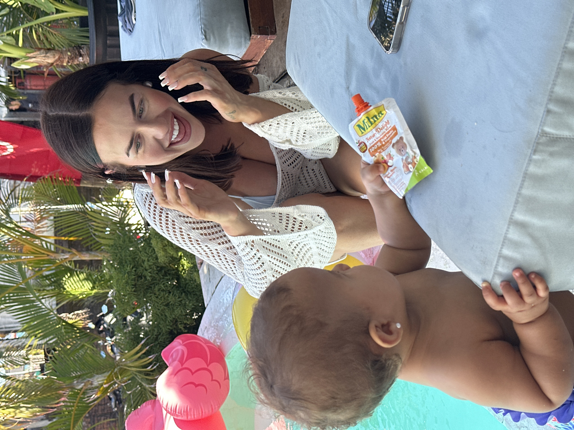 A medium, eye-level shot captures a scene outdoors in what appears to be Kuta, Indonesia, given the tropical foliage. A woman with dark hair and a white crochet cover-up, who appears to be Caucasian, is seen smiling. She is interacting with a baby, also appearing Caucasian, who is holding a “Milna” brand baby food pouch. The baby seems to be in or near a body of water, possibly a pool, suggested by the blue hue and partially visible inflatable flamingo toys. There is a gray, cushioned outdoor sofa visible in the background, as well as a cell phone on the sofa. Tropical plants, specifically palm trees, surround the area.