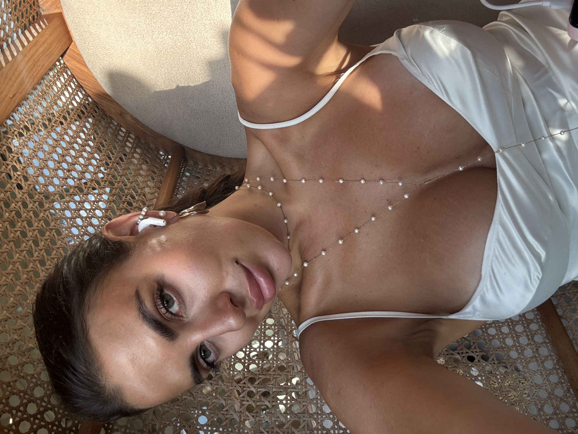 A tanned woman with dark hair and eyes is pictured in a low-angle selfie. She is wearing white wireless earbuds and a delicate pearl necklace that drapes across her chest. The woman is wearing a white dress with thin straps. She is sitting in a wooden chair with a woven wicker seat and back. The environment appears to be outdoors during the daytime, with sunlight casting shadows in the scene. While the precise location is not discernible from the image, the prompt mentions the city of North Kuta, Indonesia.