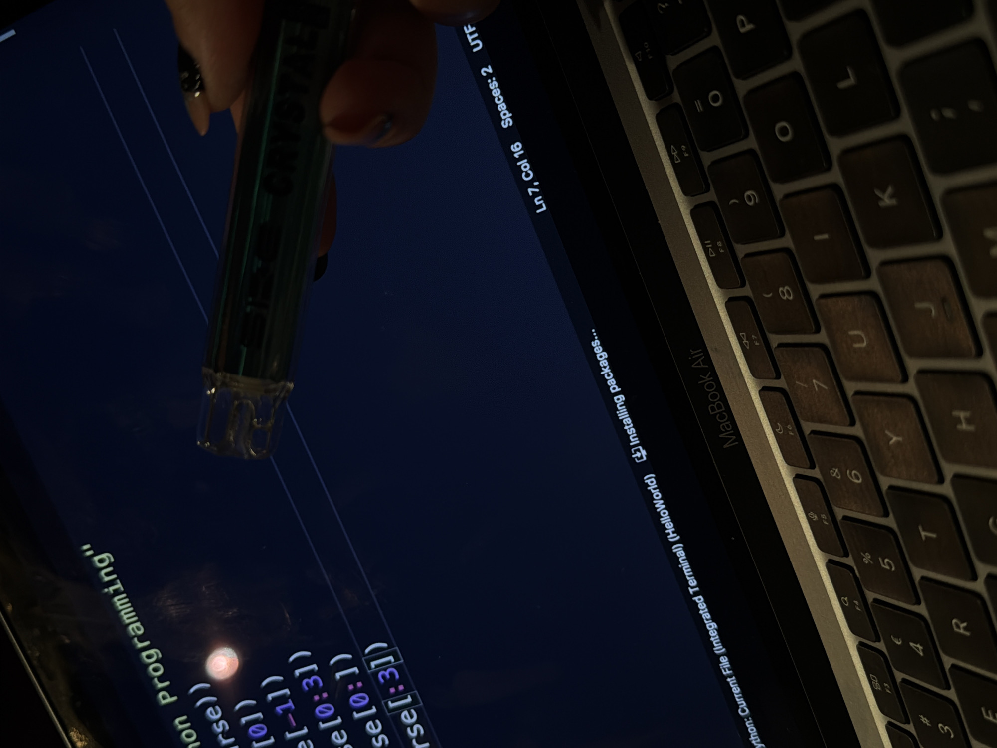 The image shows a person using a stylus on a MacBook Air. The laptop displays code, specifically Python programming, within an integrated terminal. The status bar indicates that the current file is "HelloWorld" and the system is installing packages. The cursor is located on line 7, column 16, with a setting of 2 spaces using UTF encoding. The MacBook Air's keyboard is partially visible on the right side of the frame. The location is impossible to determine based solely on the image. There is no evidence suggesting the scene is located in Plovdiv, Bulgaria, or any other specific city.