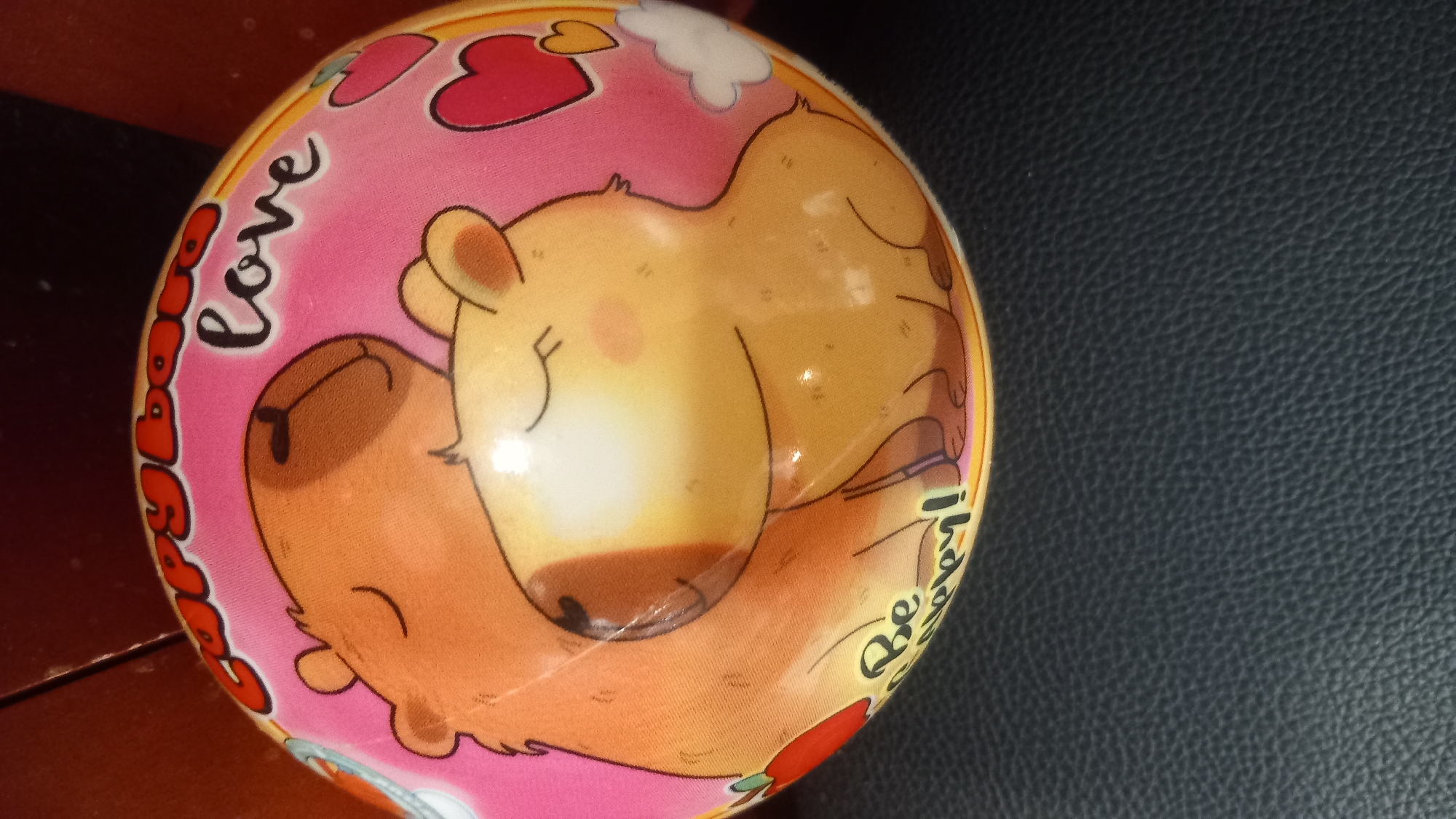 The image shows a round object with a cartoon illustration of two capybaras. The capybaras are depicted in a loving embrace, with closed eyes and blushing cheeks. The background is pink and features the words "Capybara love" and "Be happy!". There are also small heart shapes and a cloud. The object appears to be a ball or a stuffed toy. The object is resting on a dark, textured surface, possibly leather. The top edge of the object is partially obscured by a darker, wooden surface. There is no discernible connection to the city of Kos, Greece.