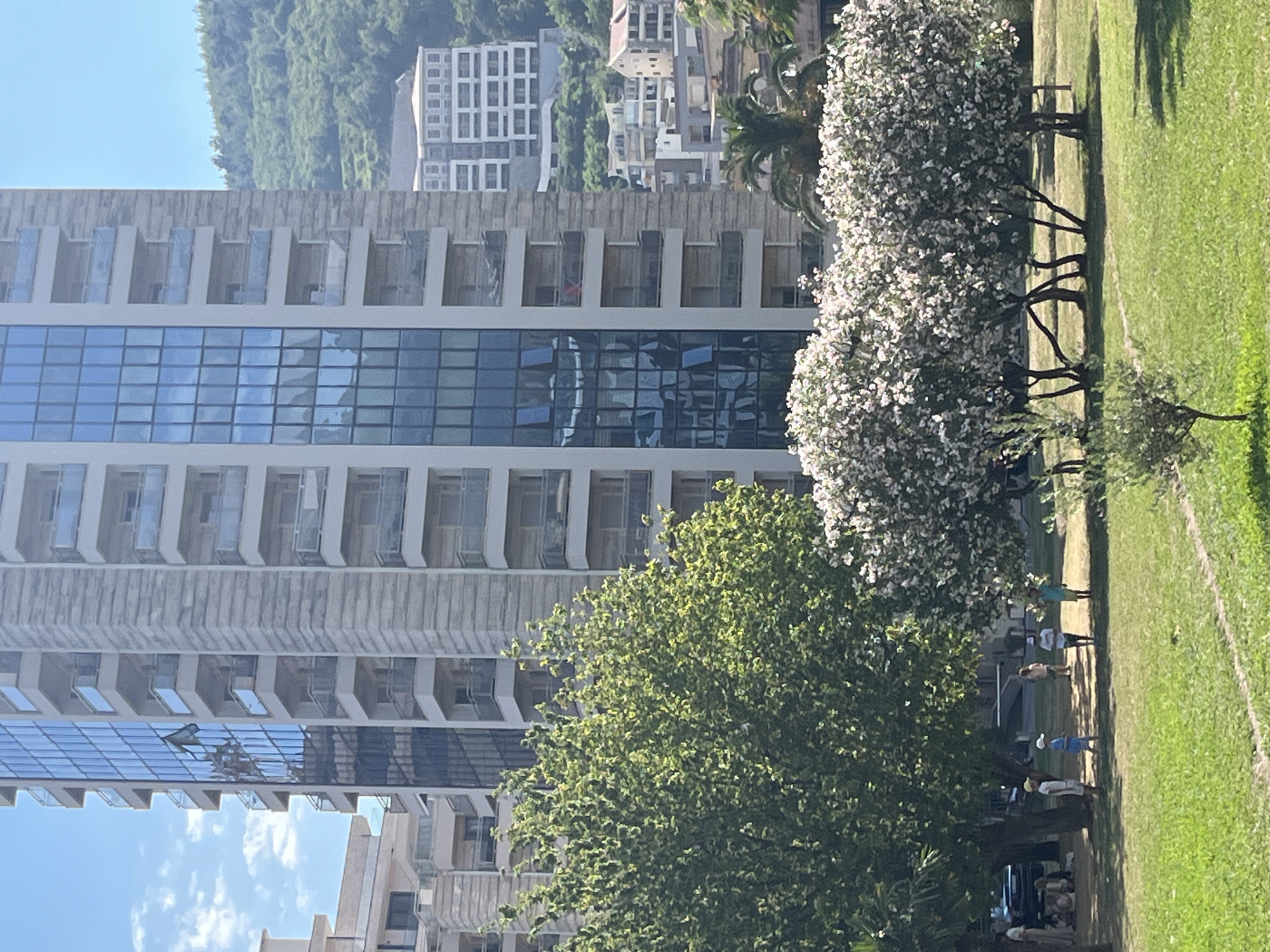 This image captures a scene in the city of Budva, Montenegro. In the foreground, a grassy lawn is visible with a few small trees, one with sparse green leaves and another with abundant white flowers. Beyond the lawn, several children are scattered, some standing and observing, while others are seated. A group of adults are also present in the background, near a parked car.
Dominating the mid-ground and background is a tall, modern apartment building with a facade of light-colored stone and a central section of blue-tinted glass windows. Many of the windows on the glass section are open. Balconies with glass railings extend from the building. To the right of this building, a lush green, mountainous landscape rises, dotted with trees and other buildings. Another multi-story building with a white facade and numerous windows is situated further up the hillside, and closer to the foreground, a cluster of smaller residential buildings with red-tiled roofs is visible. The sky is blue with scattered white clouds.