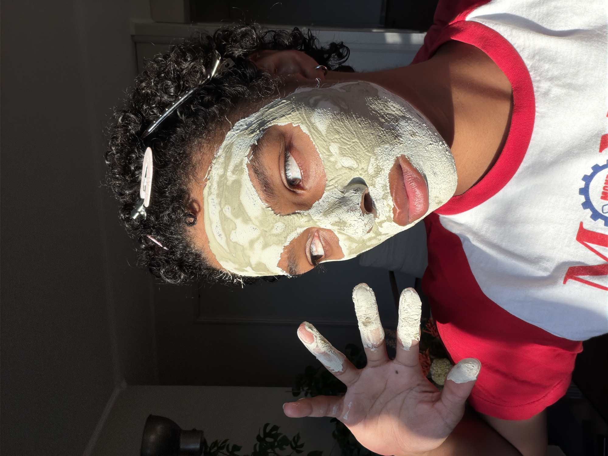 A person with curly dark hair, held back by several hair clips, is indoors, with their face partially covered in a light green face mask. They are wearing a red and white raglan t-shirt. Their left hand is raised, with fingers also covered in the face mask. The lighting suggests it is daytime, with sunlight casting shadows and illuminating parts of the scene. A plant is visible in the background to the left, and a doorway or hallway is visible to the right. The overall setting appears to be a domestic indoor environment. The location in Champs-sur-Marne, France is not discernible from the image.