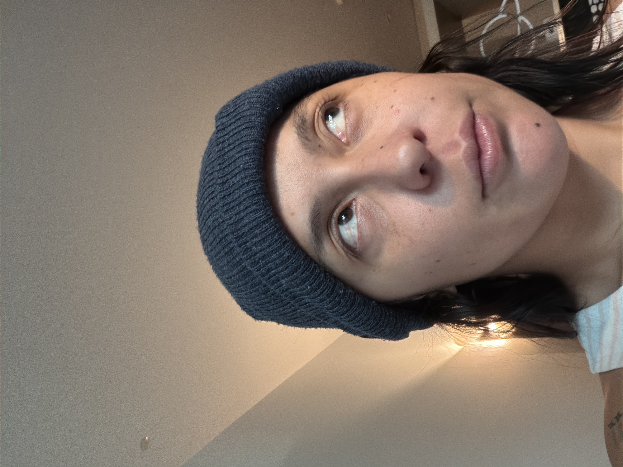 A selfie of a person wearing a dark blue beanie is angled upwards. The person has light skin, brown eyes, and dark hair. The background is a plain wall. It is not possible to tell from the image if the location is L'Hospitalet De Llobregat, Spain.