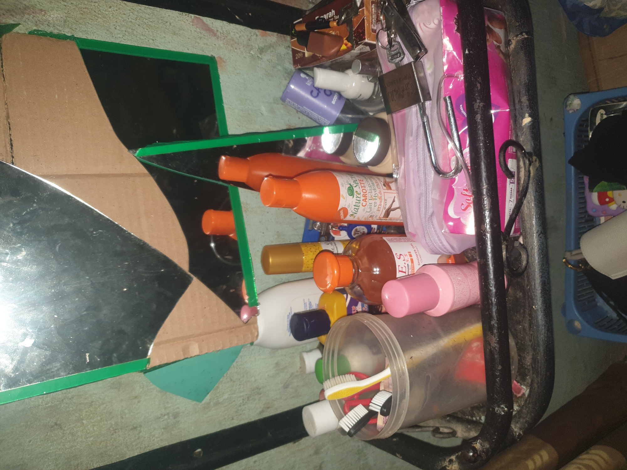 The image depicts a cluttered shelf or storage area with various personal care products and a broken mirror. In the foreground, a transparent plastic container holds several toothbrushes with black and white bristles, along with a yellow and white toothbrush. Next to the container are bottles of lotion or soap, some with orange caps. A pink bottle labeled "E.S. SHOWER GEL" is also visible. A pale purple cylindrical object with a white cap is positioned behind the toothbrushes. A portion of a pale pink cosmetic bag or pouch is in the center, with a metal lock and keys resting on top. To the right, a white bottle with a blue cap is partially obscured. Further back, a white bottle with a dark cap is present. A golden-colored spray can is also in the background. A prominent feature is the large, cracked mirror with a green border, reflecting a dark environment and some of the items on the shelf. The background appears to be a rough, textured wall. The overall scene suggests a personal vanity or storage area that is in disarray.