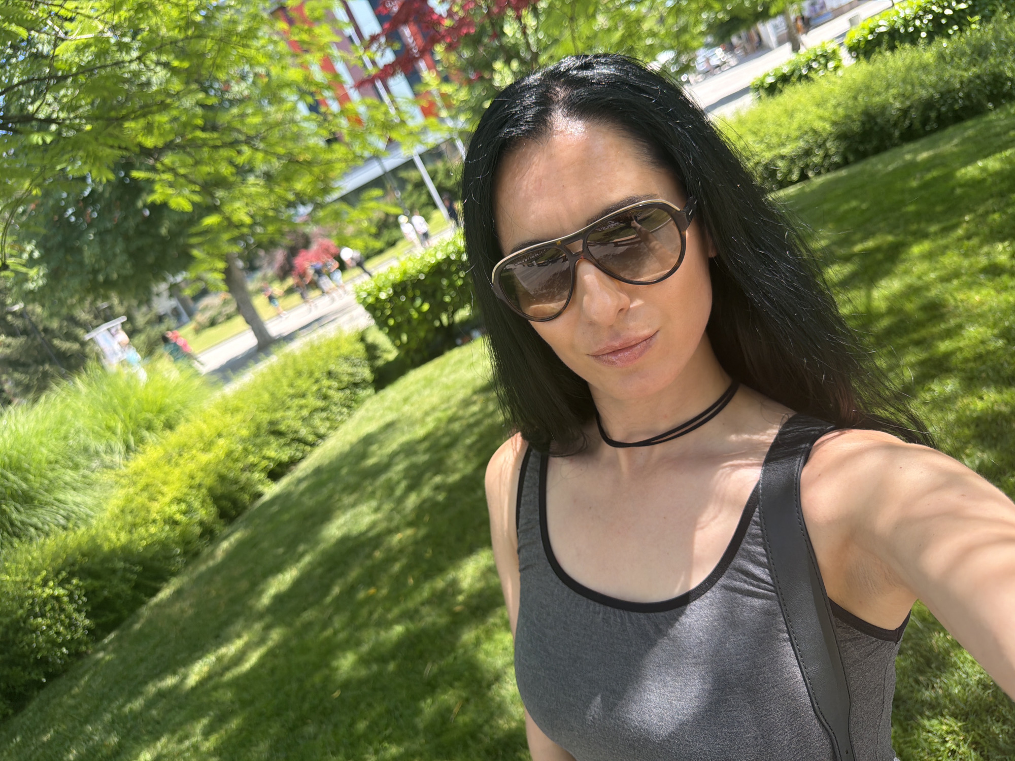 A woman with long black hair and sunglasses is taking a selfie in a park. She is wearing a gray tank top and a black choker necklace. The park has green grass, trees, and bushes. In the background, there is a building with red accents and people walking. The image is taken in the city of Pleven, Bulgaria.