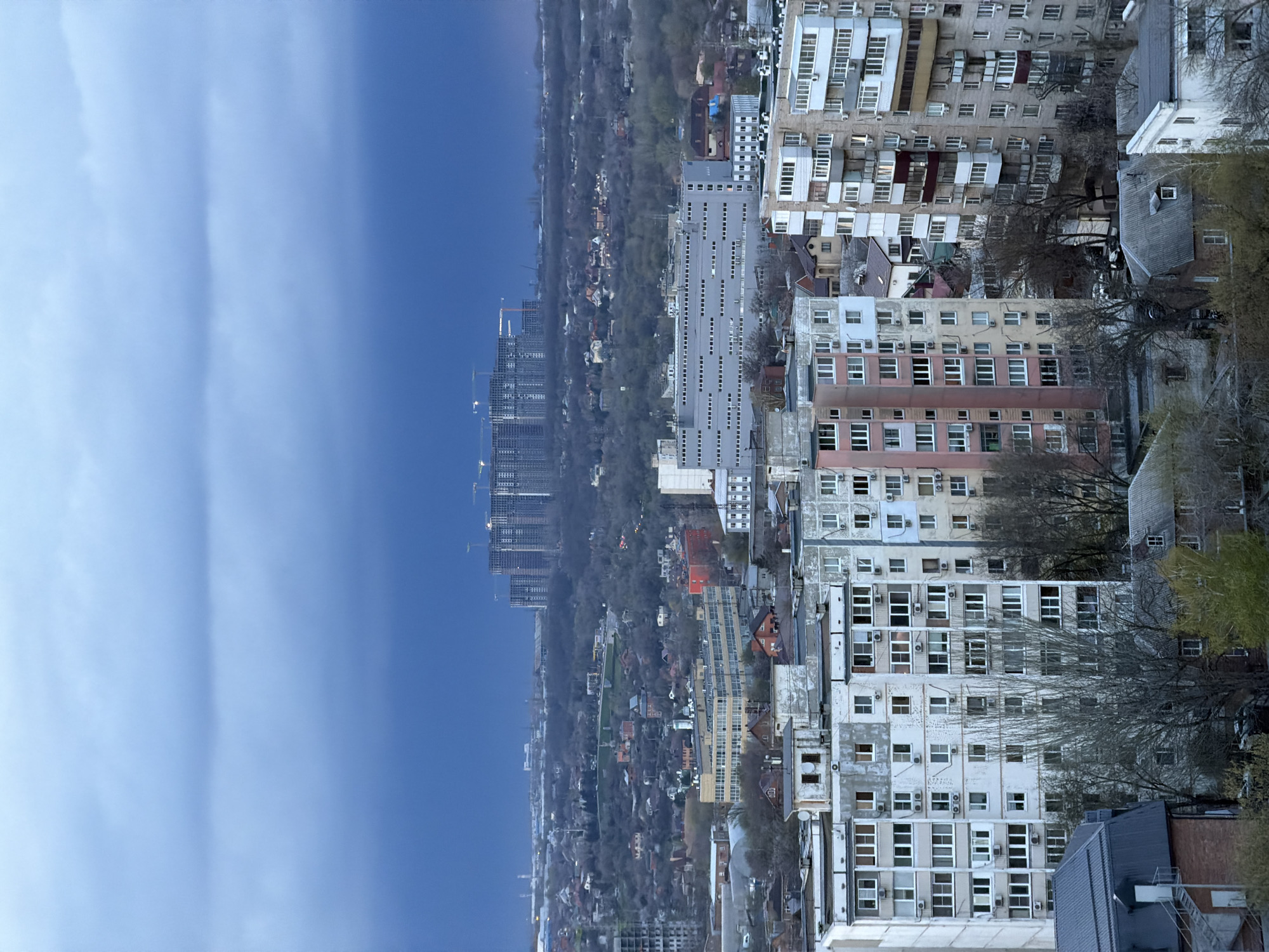 The image is an elevated, panoramic view of a cityscape in Rostov-on-Don, Russia. The composition is vertically oriented, and the perspective is likely from a high-rise building looking out over the surrounding area.
On the right side of the image, there are several multi-story residential buildings. These buildings appear to be of Soviet-era construction, with rectangular shapes and rows of identical windows. There are some trees among these buildings.
The center of the image features a green space with scattered trees and some single-family homes. This area forms a transition between the dense urban environment and the more open space further in the distance.
In the distance on the left side of the image, there are several high-rise buildings under construction. Cranes are visible, indicating ongoing building activity. The sky above is overcast with a grey tone, suggesting a cloudy or rainy day.