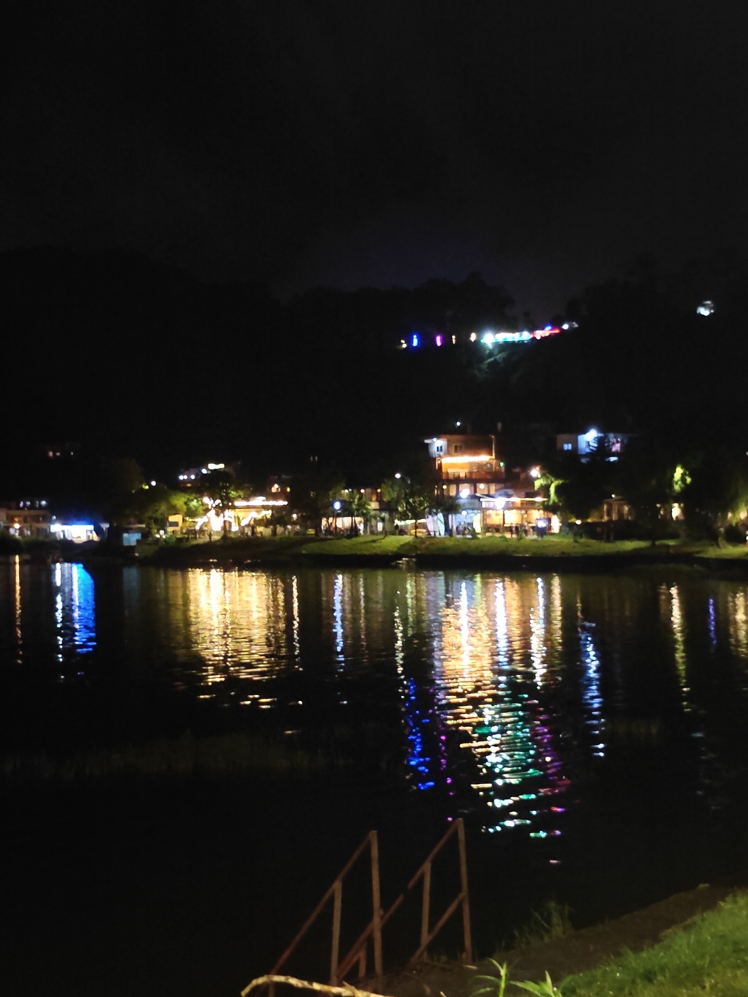 The image depicts a nighttime scene of the lakeside in Pokhara, Nepal, specifically the area along Phewa Lake, likely near Lakeside road. In the background, a hillside is visible, dotted with various buildings and illuminated by colorful lights. These lights suggest a lively commercial and residential area, possibly restaurants, hotels, or shops. The lights are reflected in the dark water of Phewa Lake, creating a vibrant, shimmering effect. In the foreground, there is a grassy bank with a wooden railing leading down towards the water, indicating a public access point or a walkway. The overall atmosphere is serene and picturesque, showcasing the beauty of Pokhara at night. There are no discernible individuals or specific actions captured in the frame.