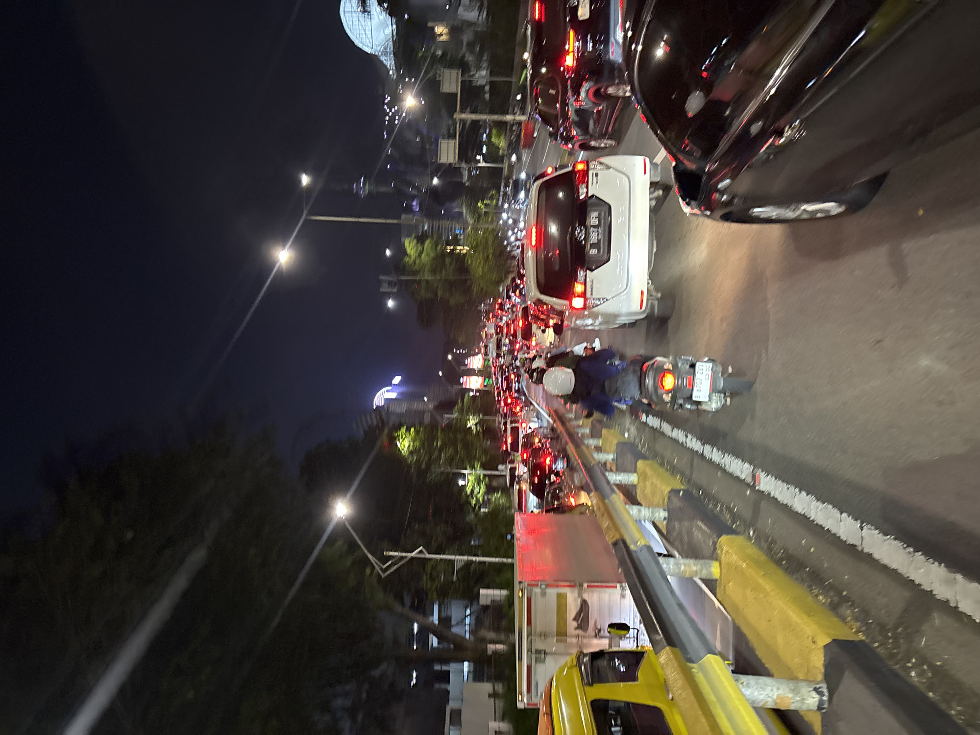The image presents a nighttime scene of a congested urban street in Central Jakarta, Indonesia. The perspective is slightly elevated, looking along a multi-lane road densely packed with vehicles, indicating heavy traffic or a significant traffic jam.
Numerous cars and motorbikes fill the roadway. In the mid-ground, a white SUV with the license plate "B 7092 UFM" is prominent, its red taillights glowing. A dark-colored car is partially visible to its right. Further down the road, a long line of vehicles extends into the distance, identifiable by their illuminated headlights and taillights. A person wearing a helmet is seen riding a motorbike, navigating through the traffic behind the white SUV.
In the immediate foreground and along the right edge, a yellow and black road barrier separates the viewer's position from the main traffic lanes. A partial view of a yellow truck or commercial vehicle is visible at the bottom right, suggesting the vantage point.
The dark sky confirms it is nighttime, and the weather appears clear. The scene is illuminated by multiple bright streetlights. In the background, several illuminated buildings stand out against the night sky, including one with a distinctive dome-like structure and another with vertical stripes. Trees also line parts of the road. Another motorbike's license plate, partially visible behind the barrier, reads "117 ZR2-". The image effectively captures the bustling and illuminated atmosphere of a major city experiencing traffic at night.