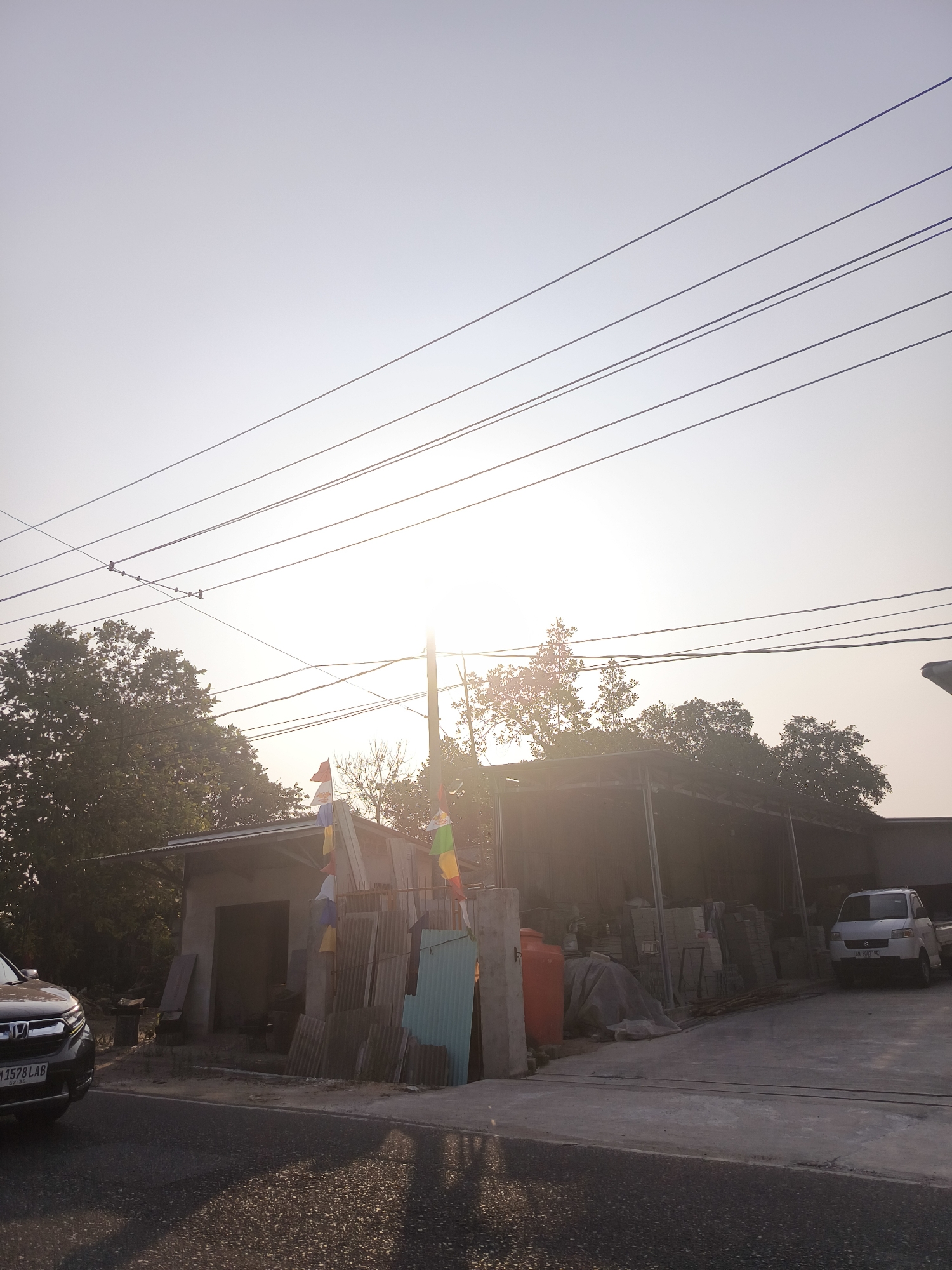 The image depicts a street scene in Pangkalpinang, Indonesia, during what appears to be sunset or sunrise, given the low angle of the sun and the warm, hazy light. In the foreground, a portion of a dark-colored SUV is visible on the left side of the frame, with its license plate reading "1578 LAB". The road in front of the vehicles is paved and shows reflections of the sunlight.
Behind the road is a low-lying building with a corrugated metal roof, which appears to be a workshop or storage area. Several stacks of what look like building materials, possibly cement boards or tiles, are leaning against the structure. Colorful pennants or flags are strung across the front of the building, adding a festive touch. To the right of this structure, a white pickup truck is parked in a concrete yard.
The background is dominated by a dense line of trees, with their foliage silhouetted against the bright sky. A utility pole with multiple wires stretching across the frame is a prominent feature in the upper portion of the image. The overall impression is one of a typical Indonesian commercial or industrial area, captured during a visually striking time of day.