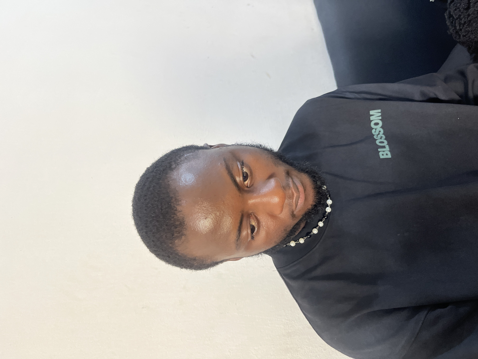 The media file is a portrait of a young Black man. He is wearing a black long-sleeved shirt with the word "BLOSSOM" printed on the sleeve in a light teal color. He is also wearing a necklace made of small pearls and black beads. The man has short, dark hair and a beard. He is looking at the camera with a slight smile. The background is a plain white wall. The lighting suggests an indoor setting, possibly with natural light coming from the side. The overall mood of the image is relaxed and casual. There are no specific cues to indicate the time of day, weather, or a precise location beyond the provided context of Gura Top, Nigeria. The image is a close-up shot, focusing on the man's face and upper body.