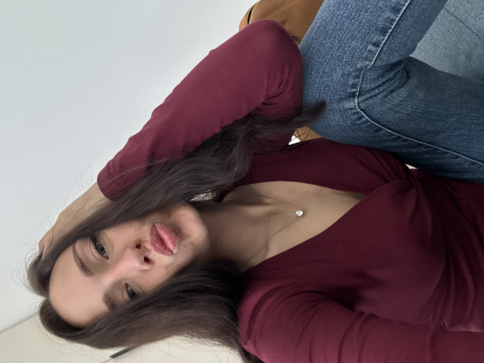 A woman wearing a maroon long-sleeved shirt, blue jeans, and a light brown boot is visible in the image. She is posed indoors against a white background. The woman has dark hair and is wearing a necklace. She is looking directly at the camera with her lips pursed. The location is not discernible from the image.