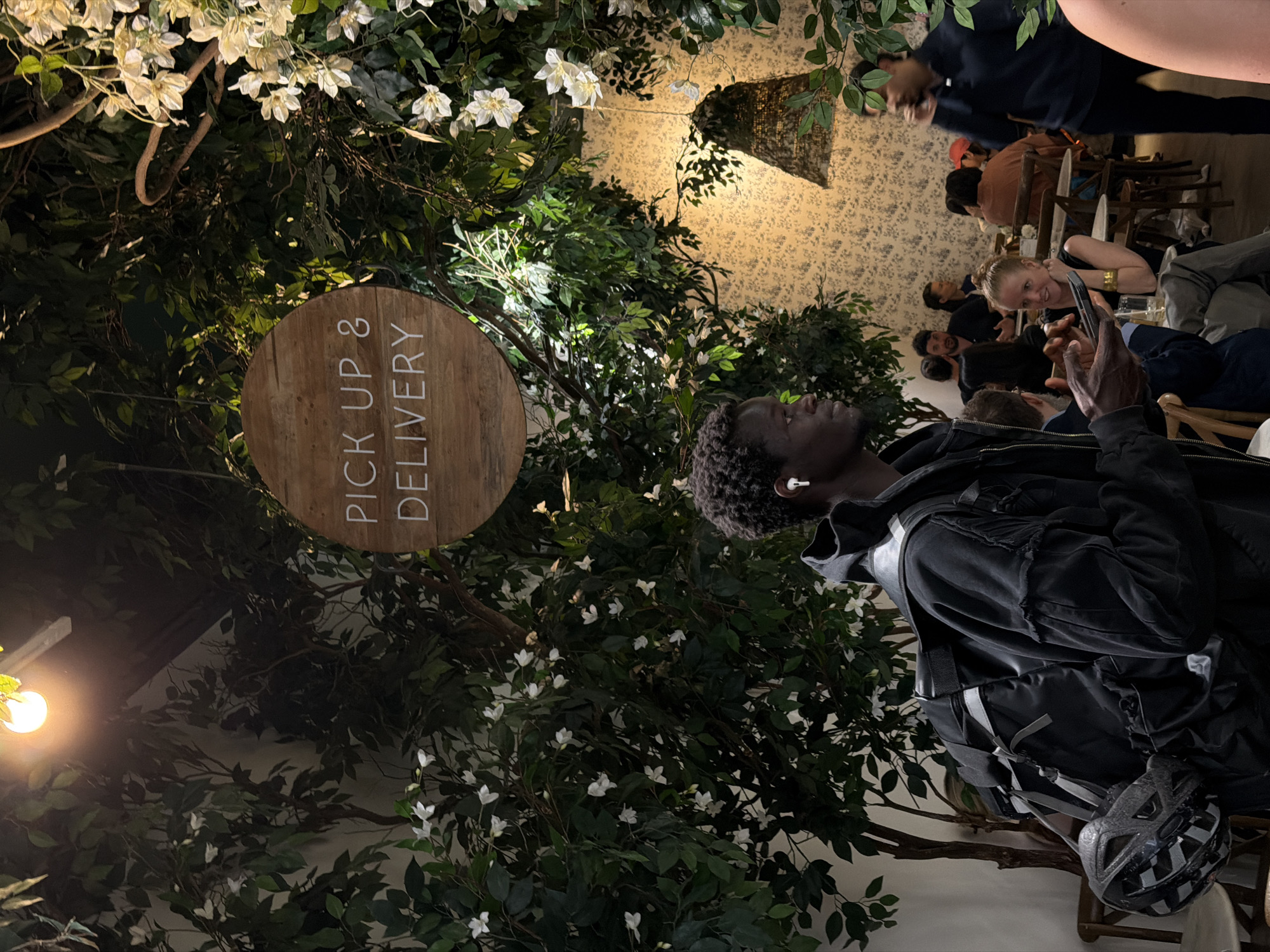 An indoor scene, likely a cafe or restaurant, is depicted with a strong emphasis on natural decor. In the foreground, a young man with dark, curly hair and dark skin stands, looking slightly to his right. He wears a dark jacket, a backpack, and white wireless earbuds, holding a smartphone in his hands. A black and white checkered bicycle helmet rests on a chair next to him.
The environment is richly adorned with lush green foliage and numerous small white flowers, suggesting an artificial indoor garden or themed aesthetic. Soft, warm artificial lighting illuminates the space, indicating it is likely evening or night. In the background, several other people are seated at tables, engaged in conversation or dining. One fair-skinned woman in the mid-ground smiles towards the viewer.
The man's attire, including the backpack and helmet, along with a prominent sign, suggests he is a delivery person waiting for an order. The atmosphere appears relaxed and inviting for patrons.
A circular wooden sign is visible to the man's left, clearly displaying the white uppercase text: "PICK UP & DELIVERY".