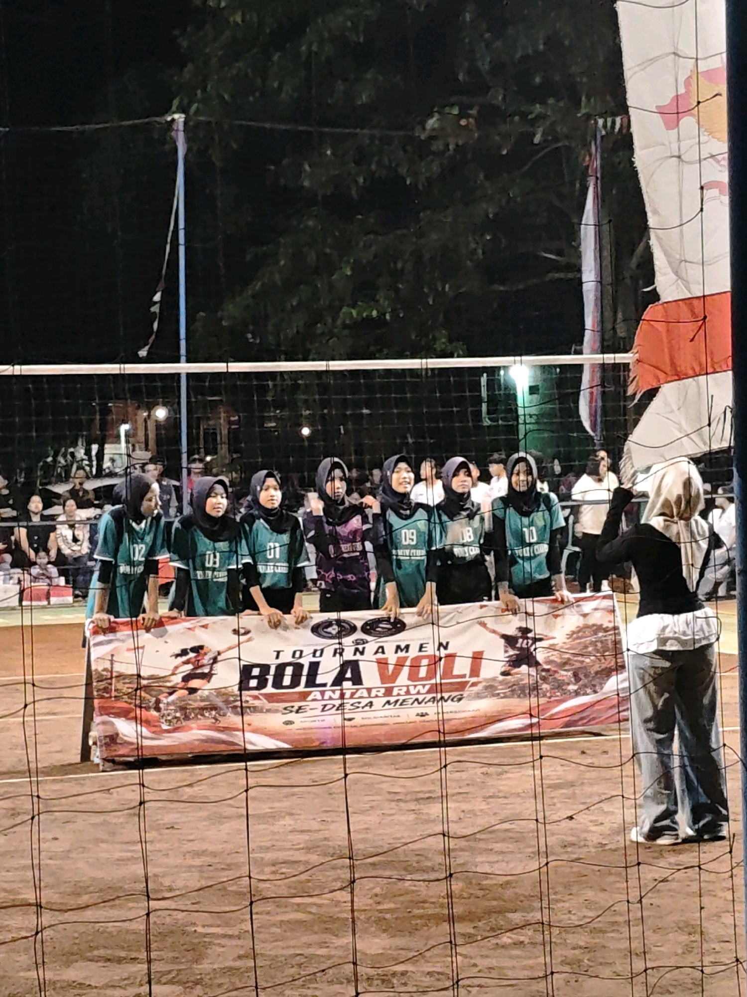 The image shows an outdoor volleyball court at night in Pagu, Indonesia. A group of young women wearing matching green jerseys and headscarves stand behind a banner that reads "TOURNAMEN BOLA VOLI ANTAR RW SE-DESA MENANG." One woman, also wearing a headscarf, stands on the other side of the net, facing the group. The court appears to be made of dirt, and a net divides the space. In the background, spectators can be seen watching the event. A flag, possibly the Indonesian flag, is visible on a pole next to the net. The lighting suggests artificial illumination, likely from floodlights.