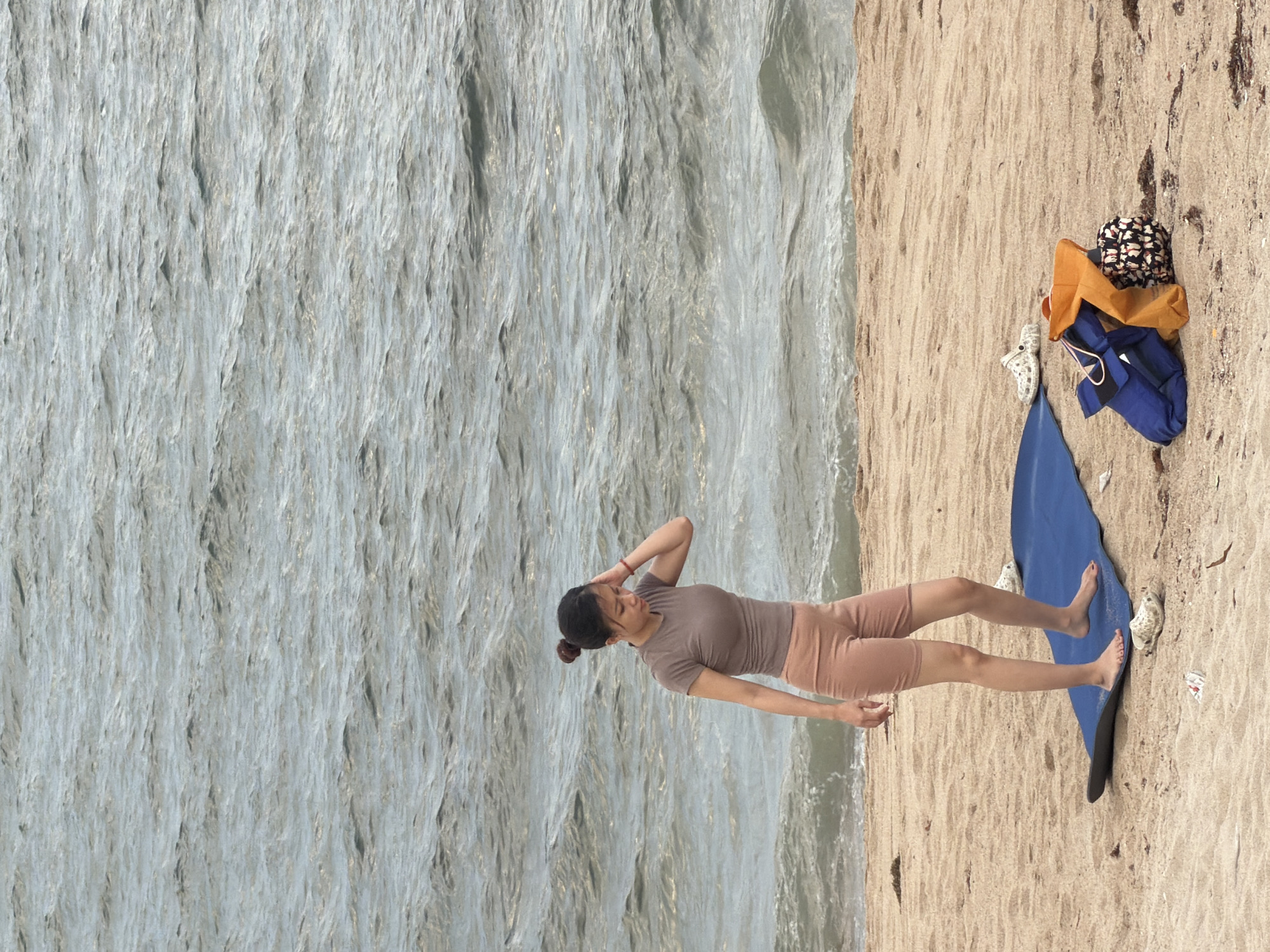 A young woman with her hair in a bun stands on a sandy beach, near the sea in Nha Trang, Vietnam. She is wearing a taupe short-sleeved shirt and taupe shorts. Her right hand is near her head, and her left arm is by her side. In front of her is a blue yoga mat. To the right of the yoga mat are a pair of white Crocs, a blue bag, an orange bag, and a patterned bag. The sea is in the background, with visible waves. The sand is light brown.