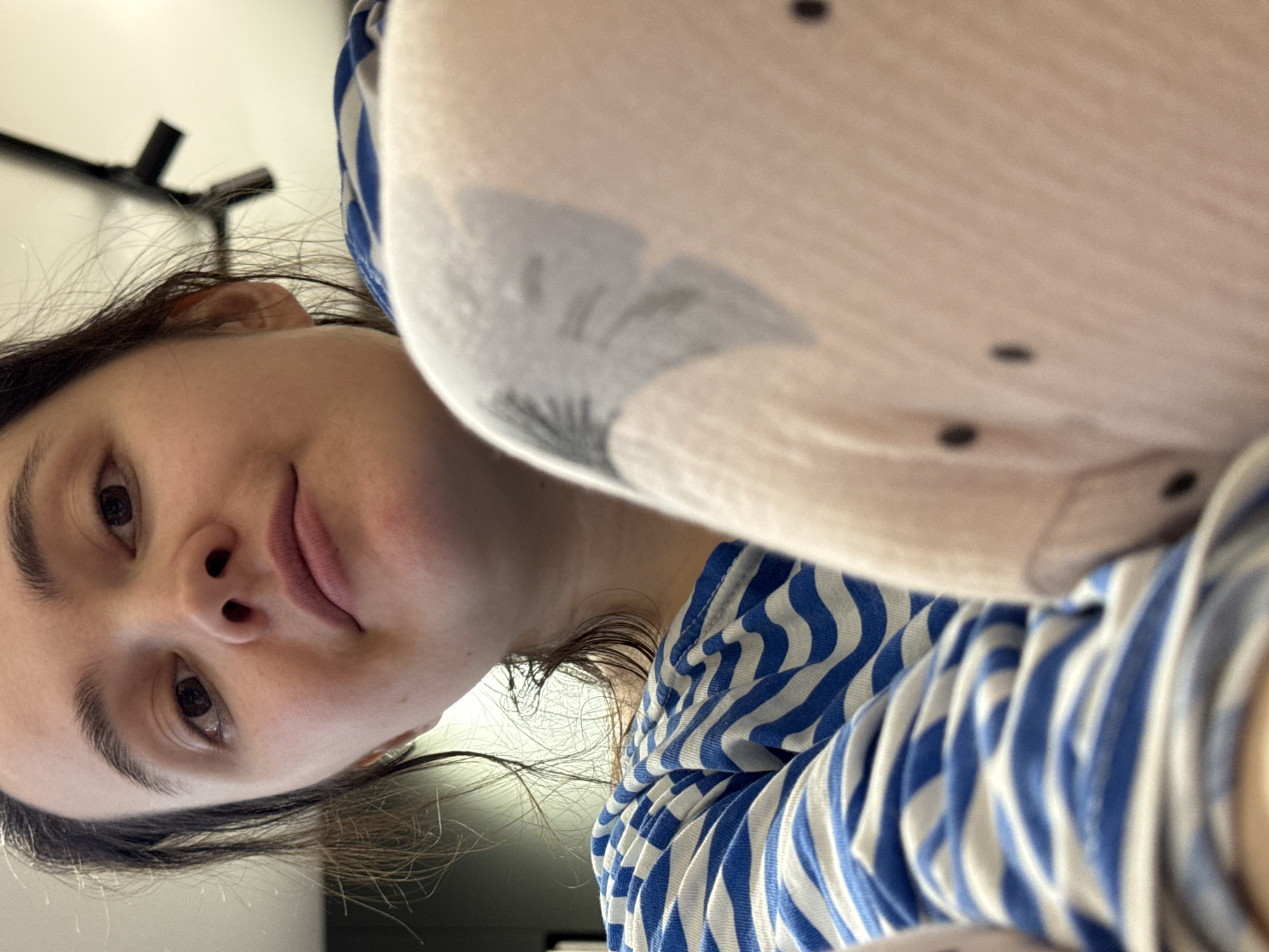 A close-up, upward-angled portrait features a young woman, likely captured in a selfie from a low perspective. Her head is tilted slightly, and she looks directly at the camera with a neutral, calm expression, her lips softly closed. She has dark hair, dark brown eyes, and prominent eyebrows. She is wearing a blue and white striped long-sleeved top, visible on her torso and arm. In the foreground, to the right and upper right, a large, blurred section of a light-colored, textured fabric item is visible. This item has small dark dots and a larger, blurry grey pattern, possibly an abstract design or a stylized animal silhouette, suggesting it could be a blanket, a plush toy, or another piece of clothing worn or held close. The background is a plain, light-colored surface, possibly a wall or ceiling, with a dark, angular structure (perhaps a light fixture or part of a furniture frame) visible in the upper left. The lighting is soft and even, indicating an indoor setting. No specific time of day or weather conditions are discernible, and there is no visible text. The location context for this image is Izhevsk, Russia.