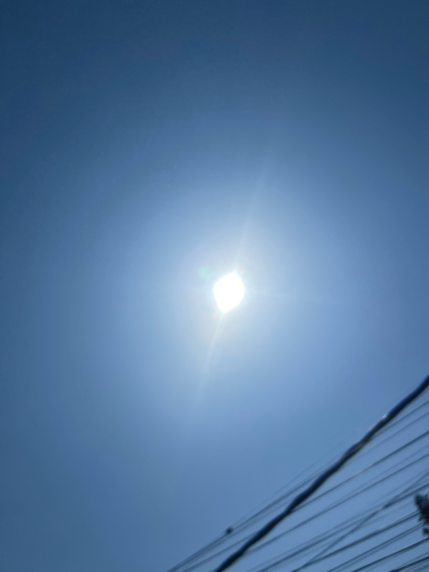 The image depicts a bright, white light source, likely the sun, centered in a clear blue sky. The light is intensely bright, creating a lens flare effect that emanates outwards. In the lower right portion of the frame, a series of dark, thin lines, consistent with power lines, are visible. The lines are out of focus and diagonally oriented. There are no people, animals, or buildings clearly visible in the frame. The scene appears to be outdoors, under a daytime sky. Without additional context, it is not possible to definitively determine the location in Denpasar, Indonesia.