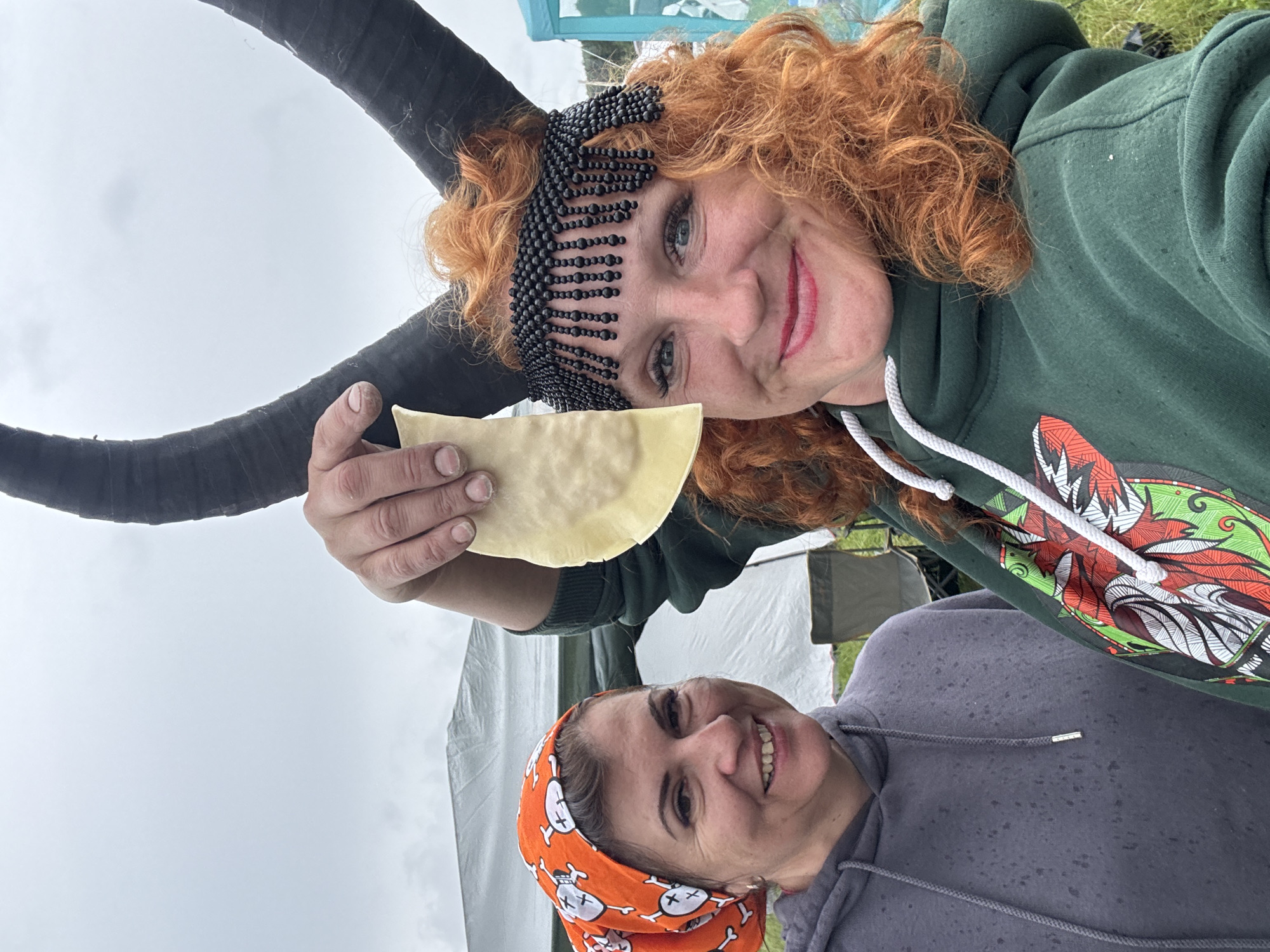 This outdoor photograph features two women smiling at the camera under an overcast sky, possibly during a festival or outdoor event in Большая Кивара, Russia.
The woman in the foreground has curly red hair and is wearing a dark green hooded sweatshirt adorned with a colorful, stylized graphic design on the right side, resembling a creature's head with sharp features and tribal patterns. She has a black beaded headpiece covering her forehead and what appear to be large, dark horns extending from behind her head. She holds up a light-colored, crescent-shaped piece of dough or pastry in her left hand, which is slightly dirty. She is smiling broadly with red lipstick.
Behind her and to the right, another woman is also smiling. She wears an orange bandana patterned with white skull and crossbones designs and a dark grey hooded sweatshirt.
The background shows hints of light-colored tents or tarps and green grassy ground, suggesting an outdoor gathering. The sky is uniformly light grey, indicating an overcast day. Both women appear happy and engaged. There is no discernible text visible in the image.