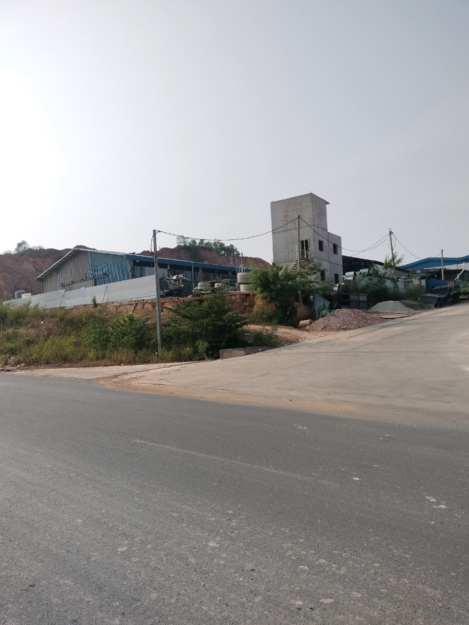 The image shows an industrial area with several buildings and a paved road. In the foreground, there is a grey asphalt road that curves to the right. Beyond the road, on a raised embankment, is a long, low-rise building with a corrugated metal roof and blue siding. To the right of this building, a taller, concrete structure with multiple stories and windows stands. Various construction materials, including piles of bricks and sand, are visible around the base of the concrete building. Further in the background, other industrial structures with blue roofs are present, along with some trees and a barren, reddish hill. The sky is clear and bright. This scene is characteristic of an industrial zone in Batam, Indonesia, possibly an area dedicated to construction or manufacturing. There are no people or animals visible in the image.