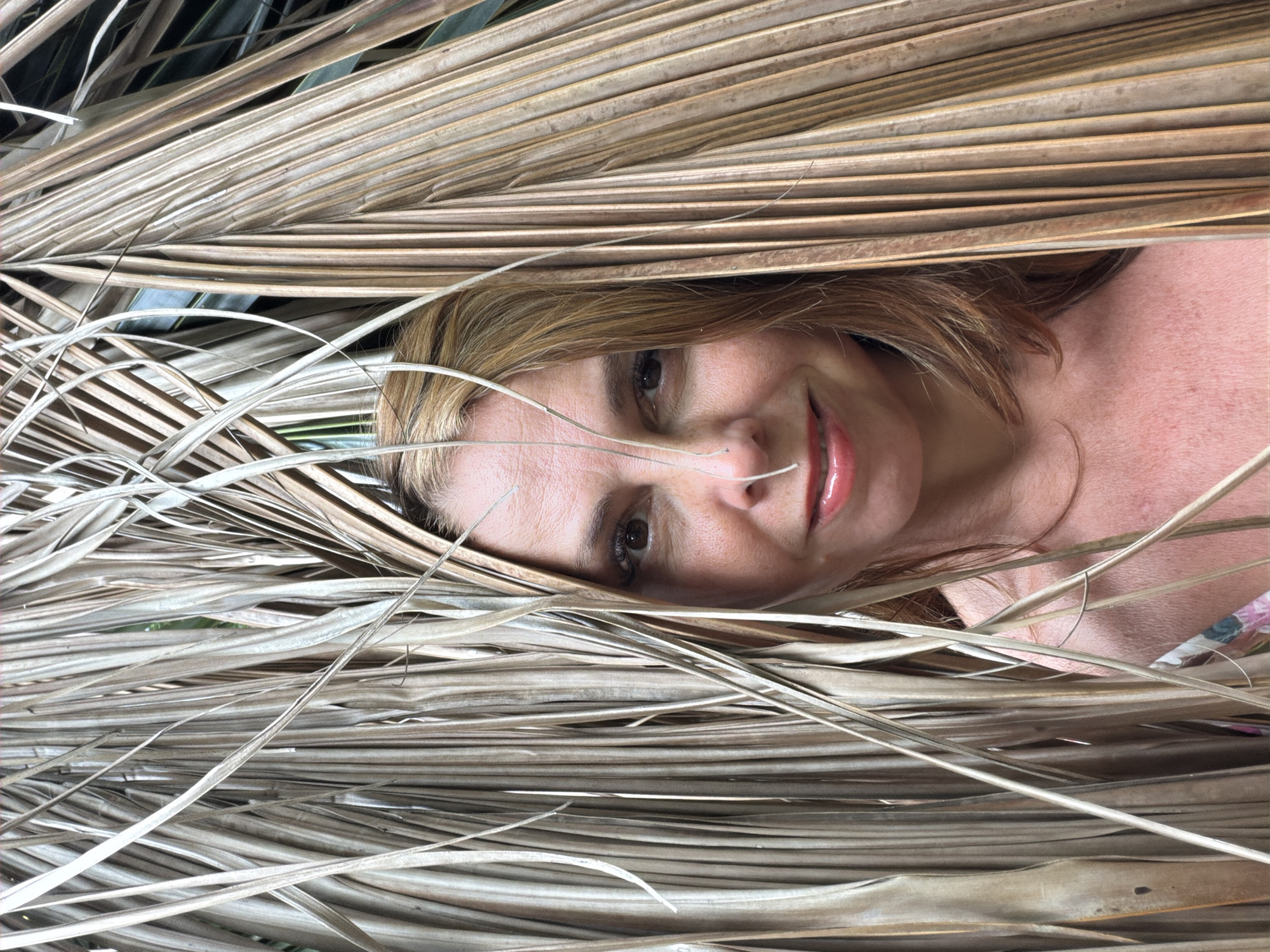 A medium close-up portrait of a fair-skinned woman with auburn hair and brown eyes, looking directly at the camera. Her face is framed by dry, brown palm fronds, which obscure parts of her body and hair. She is wearing a coral-colored top. Her skin is slightly flushed, and she has noticeable pores and fine lines. Her lips are painted with a glossy, coral-pink lipstick. The background consists of more dry palm fronds and hints of green foliage, suggesting an outdoor setting. The overall impression is that the photograph was taken in Baan Mae Nam, Thailand, likely amidst tropical vegetation.