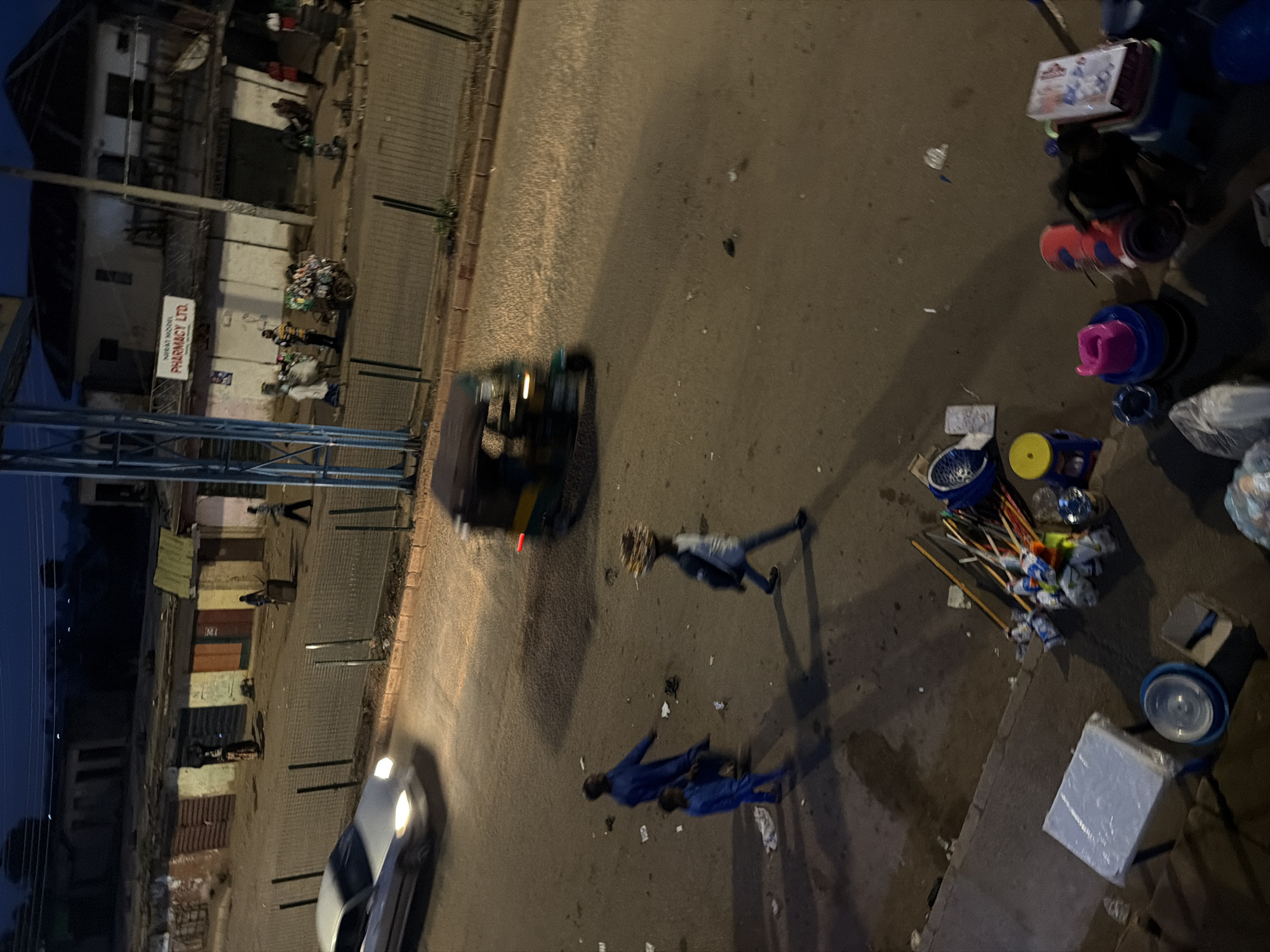 The image depicts a dimly lit street scene in Bukuru, Nigeria, likely during the evening or early morning. A silver sedan with its headlights on is in motion on the left side of the road. A three-wheeled motorized rickshaw, also in motion, is visible in the upper right quadrant of the road. Several individuals are present. Two men in blue attire are walking on the road, one taller than the other. Another individual, wearing a lighter colored shirt and dark pants, is walking across the road carrying a load on their head. Further down the street, several people are gathered near shops. One person appears to be pushing a cart loaded with goods. The environment includes several small shops lining the street, some with corrugated metal roofs. Fencing is visible along the edge of the sidewalk. On the lower right side of the image, various items are scattered on the ground, including buckets, cleaning supplies, and other assorted objects. The overall atmosphere is that of a busy marketplace or thoroughfare in a developing urban area.