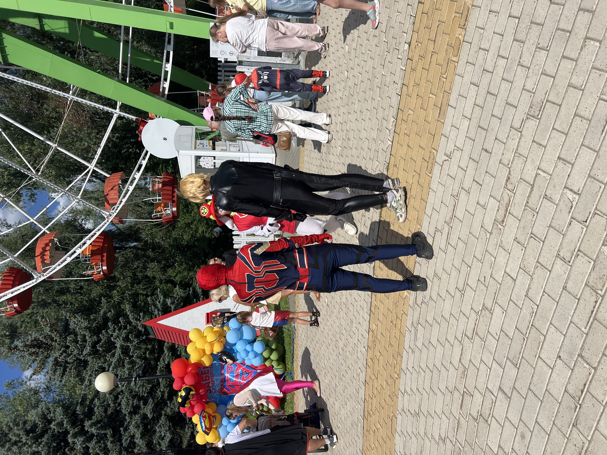 In the city of Cheboksary, Russia, at an amusement park, a group of people are gathered near a Ferris wheel. In the foreground, two individuals are dressed as superheroes. One person wears a full Spider-Man costume, while the other is in a black, form-fitting outfit resembling a comic book character. Behind them, a cluster of red, yellow, and blue balloons is arranged, with a sign that appears to say "ЗАБЕГ СУПЕРГЕРОЕВ" (Superhero Race). Several families and children are visible in the background, some looking towards the superheroes, others interacting with each other or queuing. The ground is paved with interlocking bricks. The Ferris wheel, with its red gondolas, is prominent in the upper portion of the image, set against a backdrop of green trees and a blue sky.