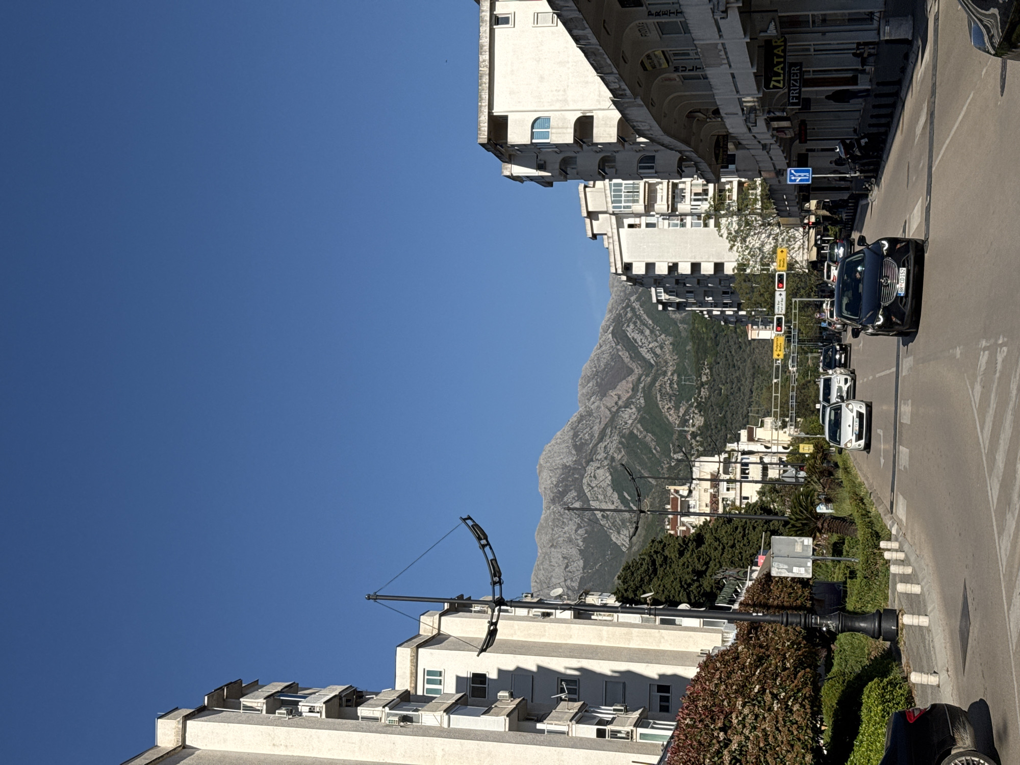 The image captures a vibrant urban scene in a town, likely Bar, Montenegro, set against a dramatic mountainous backdrop under a clear, bright blue sky. The sunny weather is evident from the distinct shadows and brilliant illumination.
In the foreground, a paved street stretches across the frame, marked with white lines for traffic and a pedestrian crossing. A black SUV, bearing the Montenegrin license plate "BR CD152" (indicating the Bar region), is stopped on the road, with other vehicles queuing behind it at a set of red traffic lights further down the street. Along the left side, a modern-looking black lamppost with an elaborate arched design stands out. The street is bordered by a green median and sidewalks, featuring various shrubs, trees (including palm trees), and concrete bollards.
Multi-story buildings, mostly light-colored and featuring numerous windows and balconies, line both sides of the street. These structures appear to house both residences and businesses. Visible text on storefront signs includes "ZLATAR" (goldsmith), "FRIZER" (hairdresser), "PARKET," and "MULTUM." Traffic signs are also visible along the roadside.
Dominating the background are large, rugged mountains, rising steeply with rocky slopes and patches of greenery, providing a striking natural contrast to the developed urban environment. The scene depicts a typical daily urban activity, with vehicles in transit or stopped at traffic signals.
