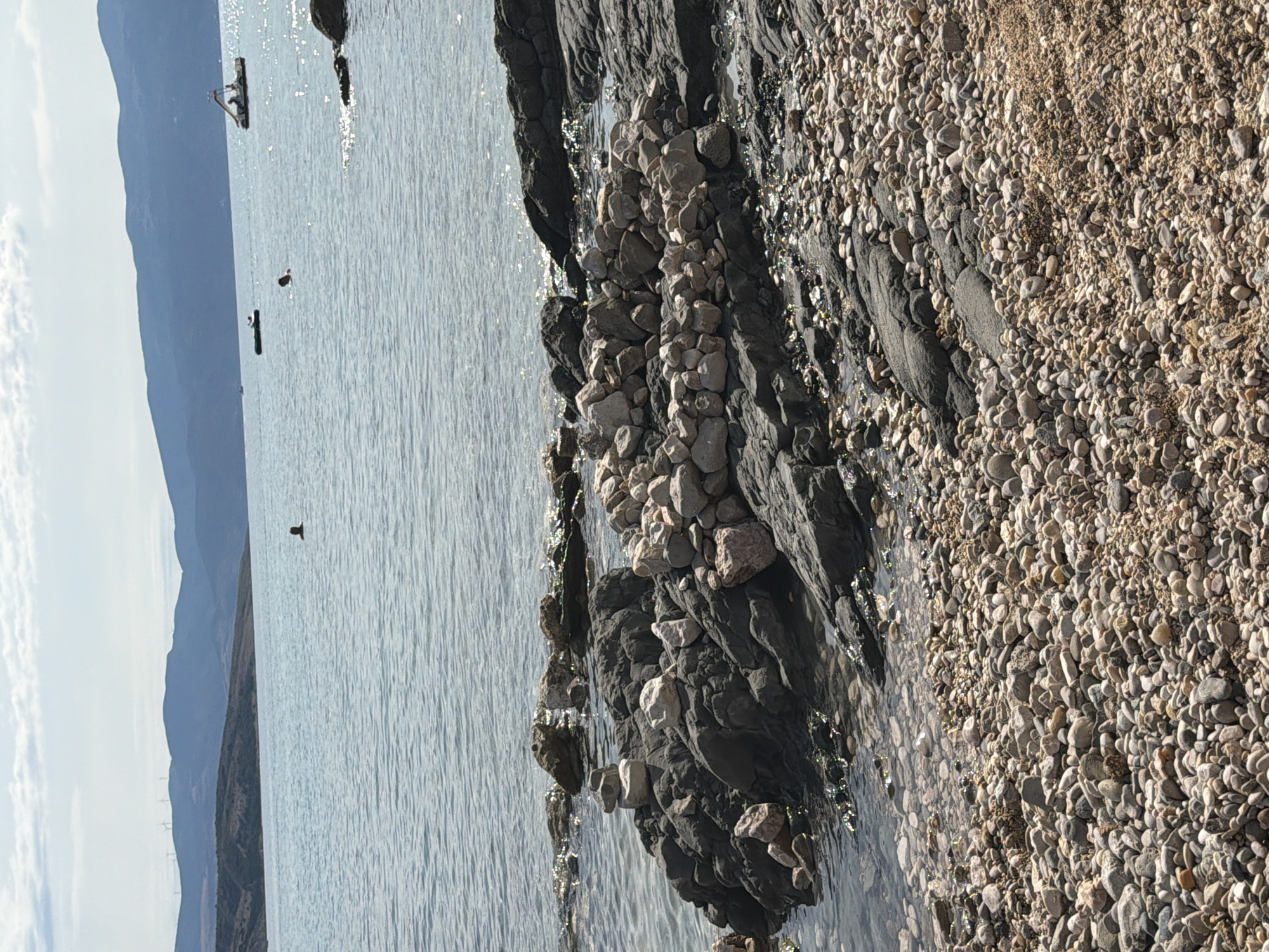 The image captures a serene coastal scene in Općina Baška, Croatia, under a partly cloudy sky. In the foreground, a rocky shore gives way to the calm, light blue sea. The shore is covered with a mix of small pebbles and sand, transitioning to larger, dark grey rocks closer to the water's edge. A circular arrangement of light-colored stones, possibly a cairn or decorative structure, is prominently placed on the rocky outcrop.
Further out in the sea, a few individuals are visible. One person is swimming, their head and shoulders above the water, while another appears to be in the distance. Several boats are also scattered across the water, including a small dinghy and a larger vessel with a structure on its deck. In the background, rolling hills with wind turbines on their crests form the horizon, beyond which lies a hazy mountain range. The water reflects the sunlight, creating shimmering highlights. The overall impression is one of a peaceful, recreational day by the Adriatic Sea.