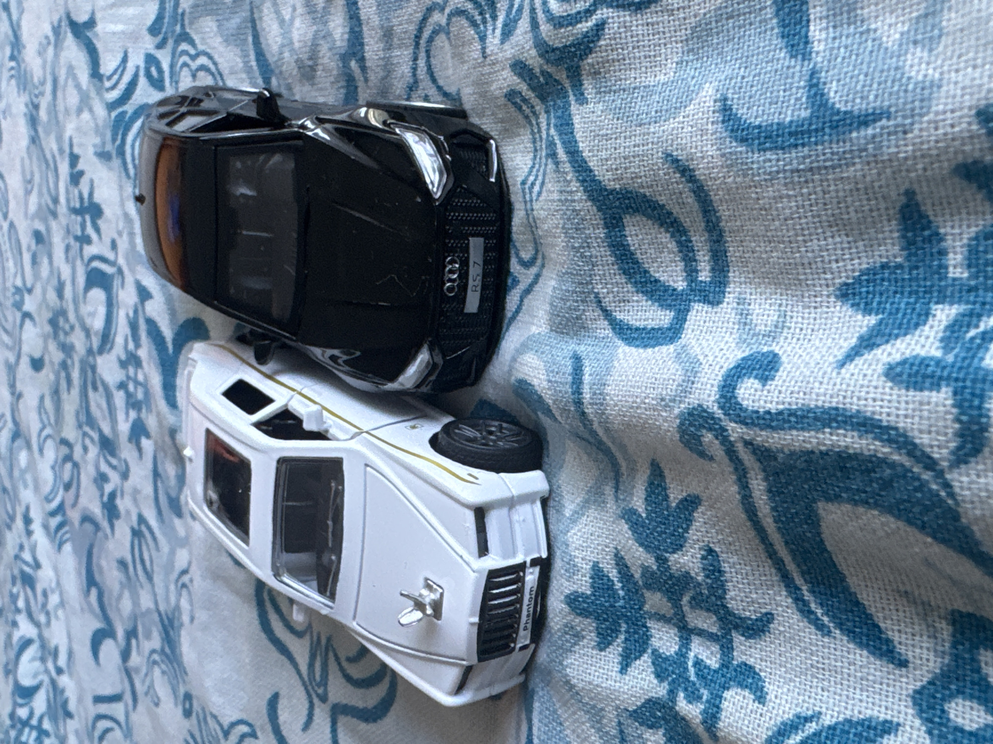 The image shows two toy cars on a blue and white patterned fabric. The top car is black and appears to be an Audi RS7, indicated by the "RS7" text and Audi logo visible on its rear. The bottom car is white and appears to be a Rolls-Royce Phantom, with "Phantom" written on its rear. Both cars are positioned on their sides, suggesting they are not in active use. The fabric has a floral or paisley pattern. The lighting suggests it is daytime. The setting appears to be indoors, on a surface like a tablecloth or bedspread. There are no people visible in the image. The overall scene is static, focusing on the toy vehicles.