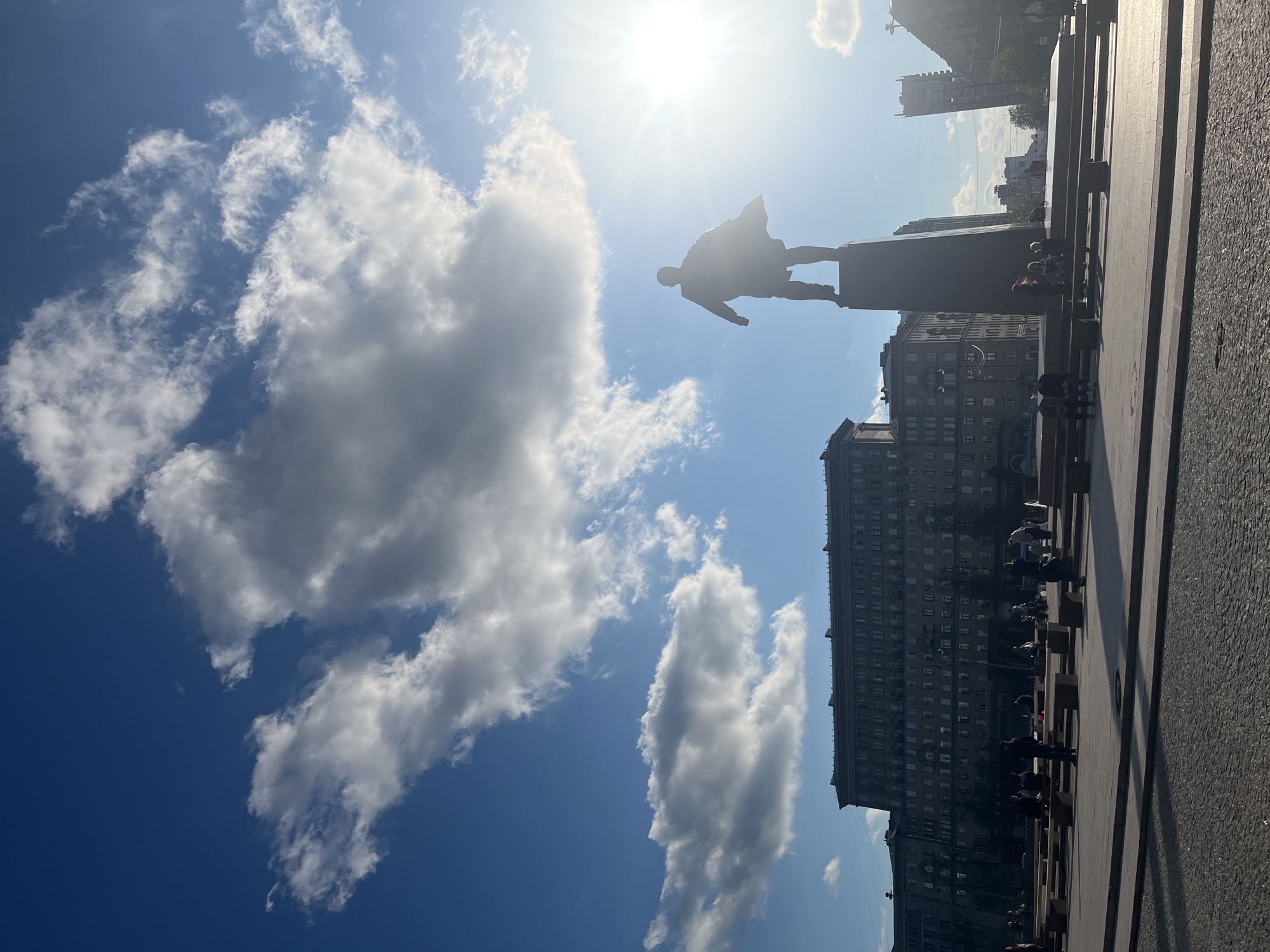 A low-angle, outdoor shot captures a statue against a backdrop of sky and clouds in Saint Petersburg, Russia. The silhouette of a male figure is atop a tall, rectangular pedestal. The sky is blue, partially obscured by puffy white clouds with sunlight shining from the top. A large apartment building stands in the background. People stand or sit on benches below the statue. The foreground shows a paved area.
