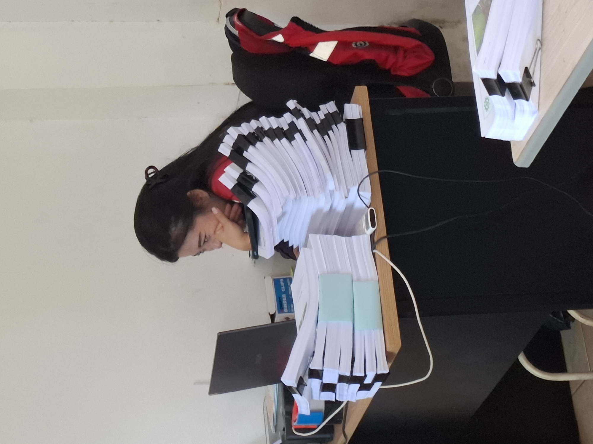 A young woman with dark hair, pulled back with a clip, is seated at a desk in an office. She is wearing a red and black striped top and has her hand resting on her chin as she looks at a laptop. In front of her are several stacks of paper, some bound with black binder clips and light blue paper bands. A printer is visible to the left of the laptop, and a red and blue taped object is on the printer. A red jacket with reflective accents is hanging on the back of her chair. Another stack of papers is on a table to the right, secured with a binder clip. The room has a plain white wall and a light-colored desk with black legs. The lighting suggests an indoor setting, possibly during daytime. The provided context places this scene in the city of Ciheulang Tonggoh, Indonesia.